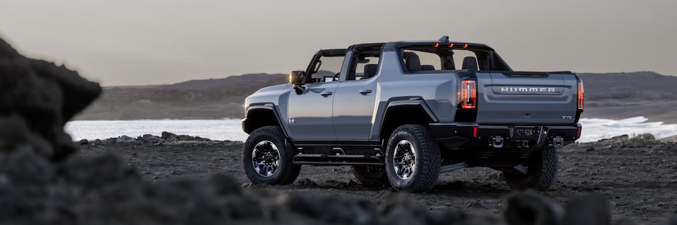 Vue latérale arrière du camion GMC Hummer EV 2025 stationné sur un chemin rocailleux avec le toit ouvert.
