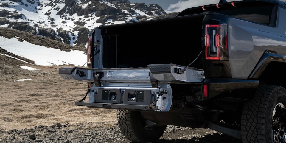 Vue arrière du hayon MultiPro ouvert du camion GMC Hummer EV 2025 devant des montagnes enneigées.