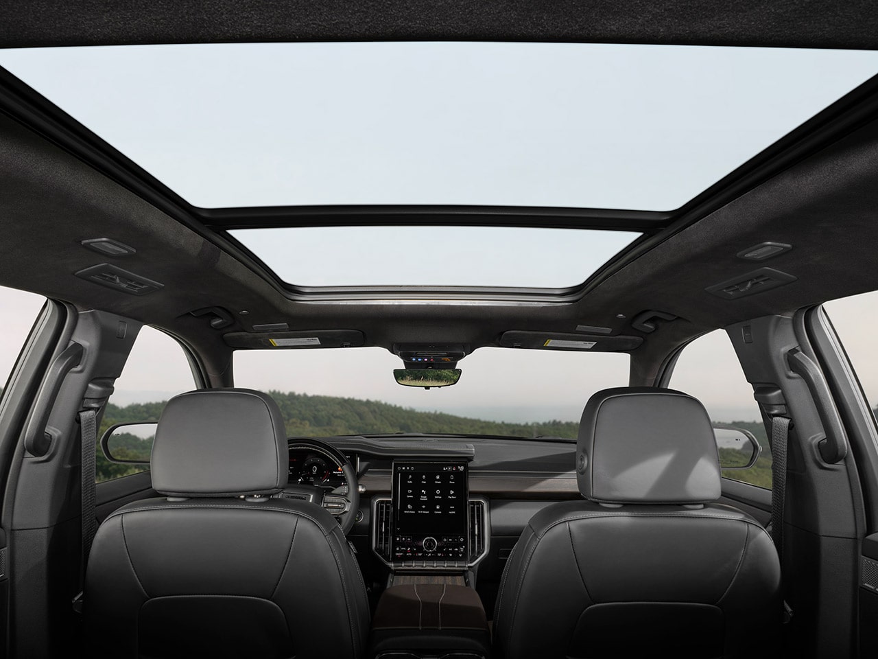 La vue intérieure d’un VUS GMC Acadia 2027 regardant vers le haut à travers le toit ouvrant panoramique, avec un paysage vert visible au loin, ainsi que le rétroviseur et l’écran d’infodivertissement.