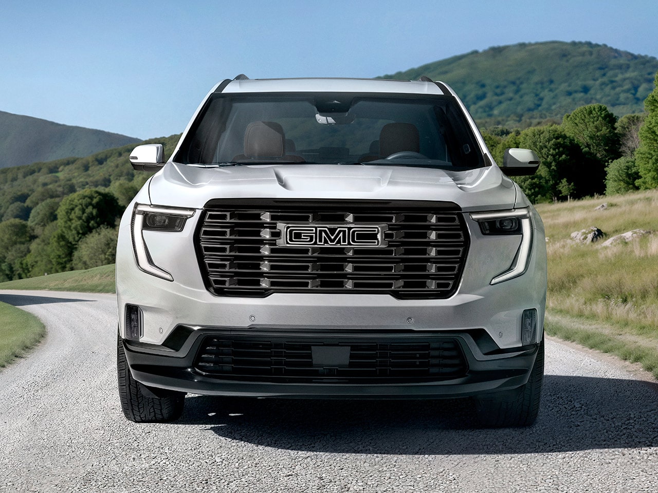 Vue de face d’un VUS GMC Acadia blanc 2027 avec le logo GMC sur la calandre, sur fond de collines ondulantes vertes.