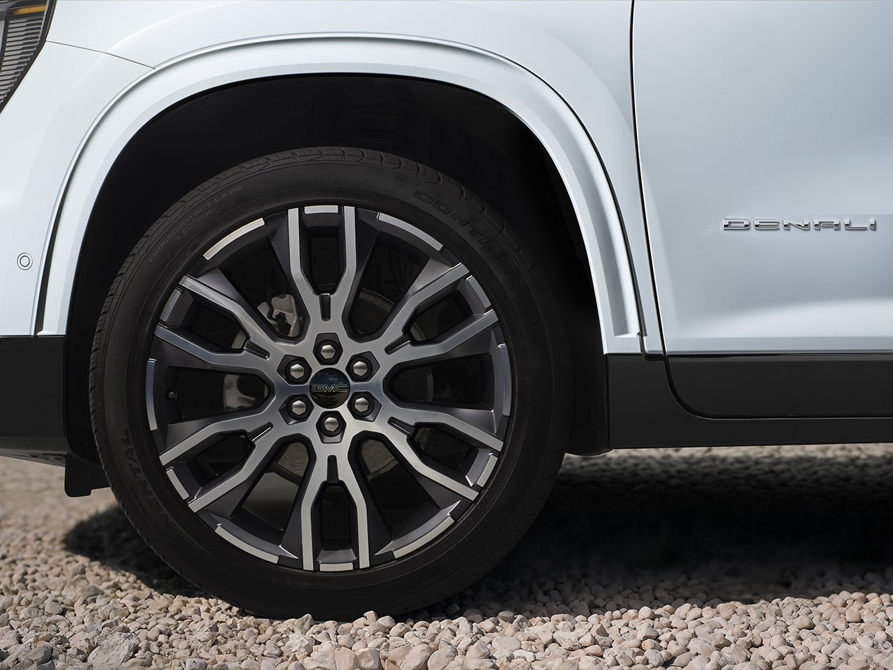 Plan rapproché d’une roue et d’un pneu à rayons multiples sur un véhicule blanc GMC Acadia Denali 2027 sur gravier.