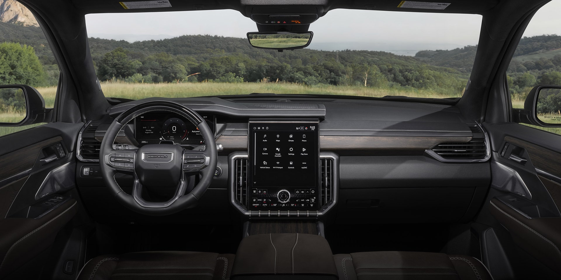 L’intérieur d’un véhicule GMC Acadia Denali 2027 montre le tableau de bord, le volant et un grand écran central avec des icônes d’applications, avec un paysage vert visible à travers le pare-brise.