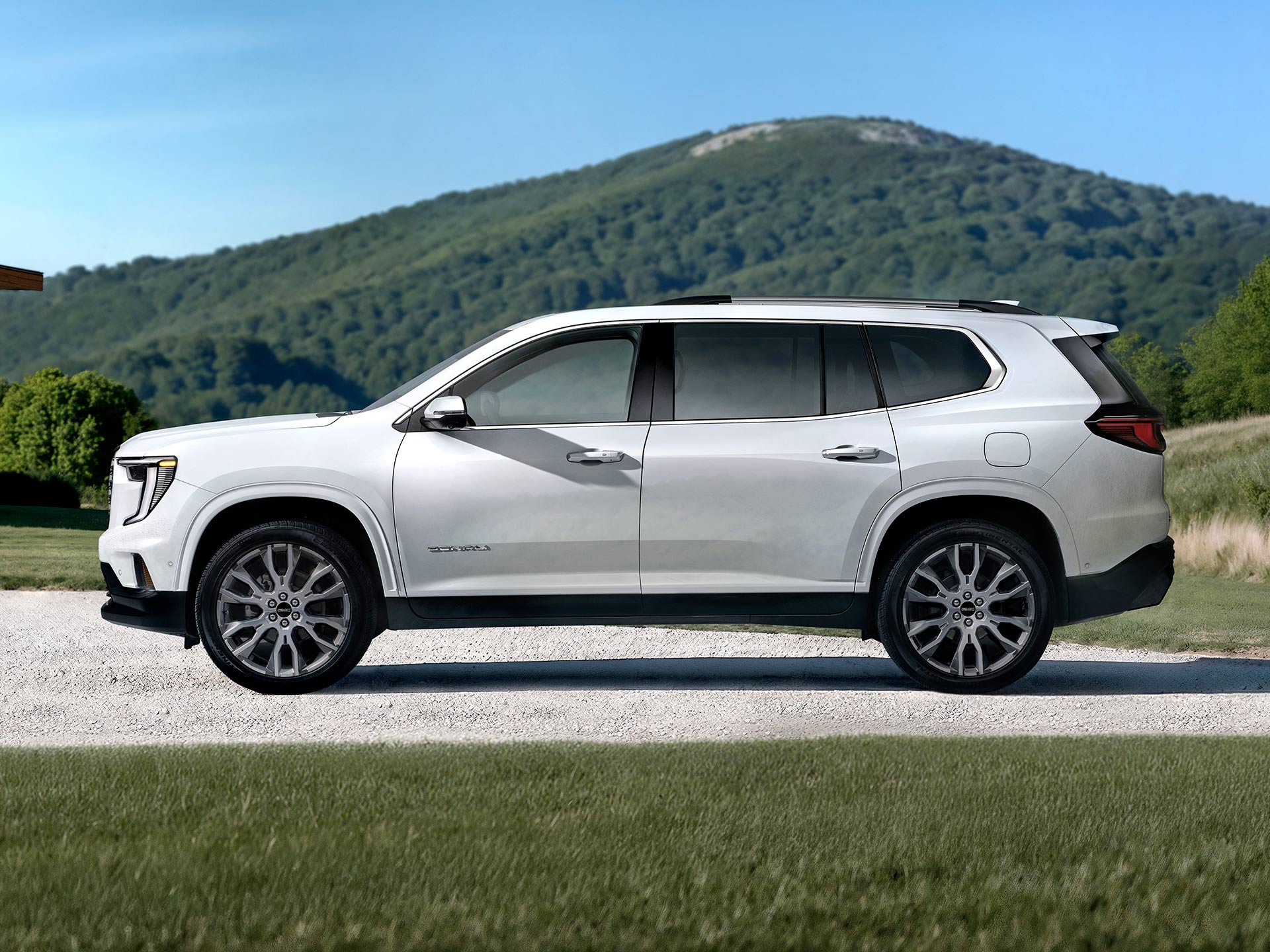 Un VUS blanc GMC Acadia Denali 2027 stationné sur une route de gravier avec des montagnes vertes en arrière-plan.