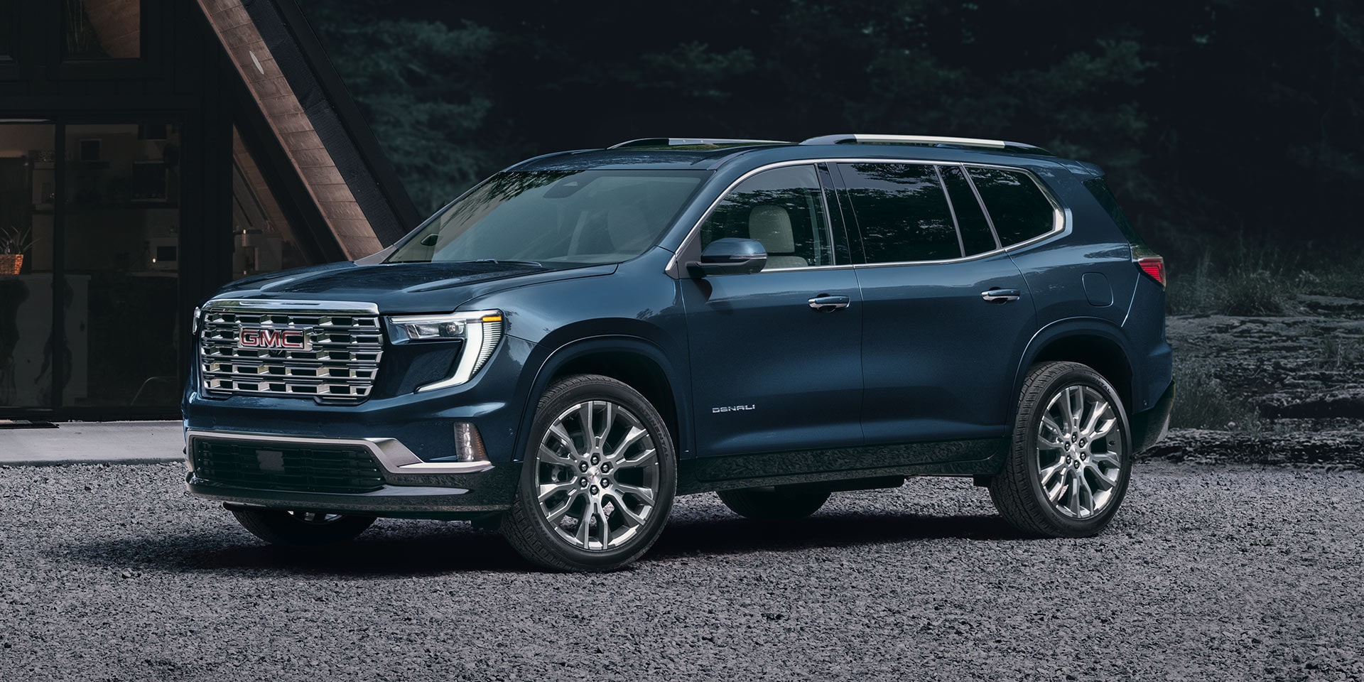 Un VUS GMC Acadia Denali 2027 bleu foncé stationné sur une surface de gravier devant une maison moderne au crépuscule.