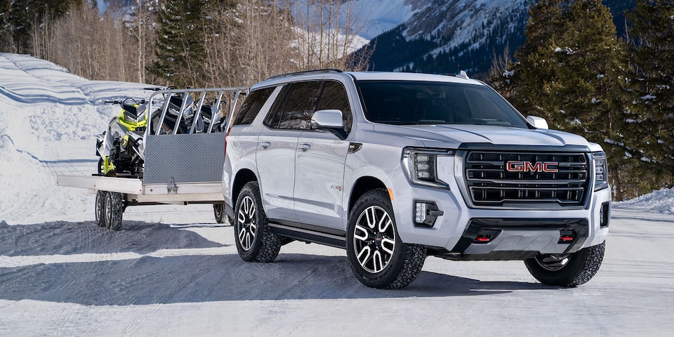 2024 GMC Yukon AT4 towing jet snowmobiles on a trailer in a snowy environment.