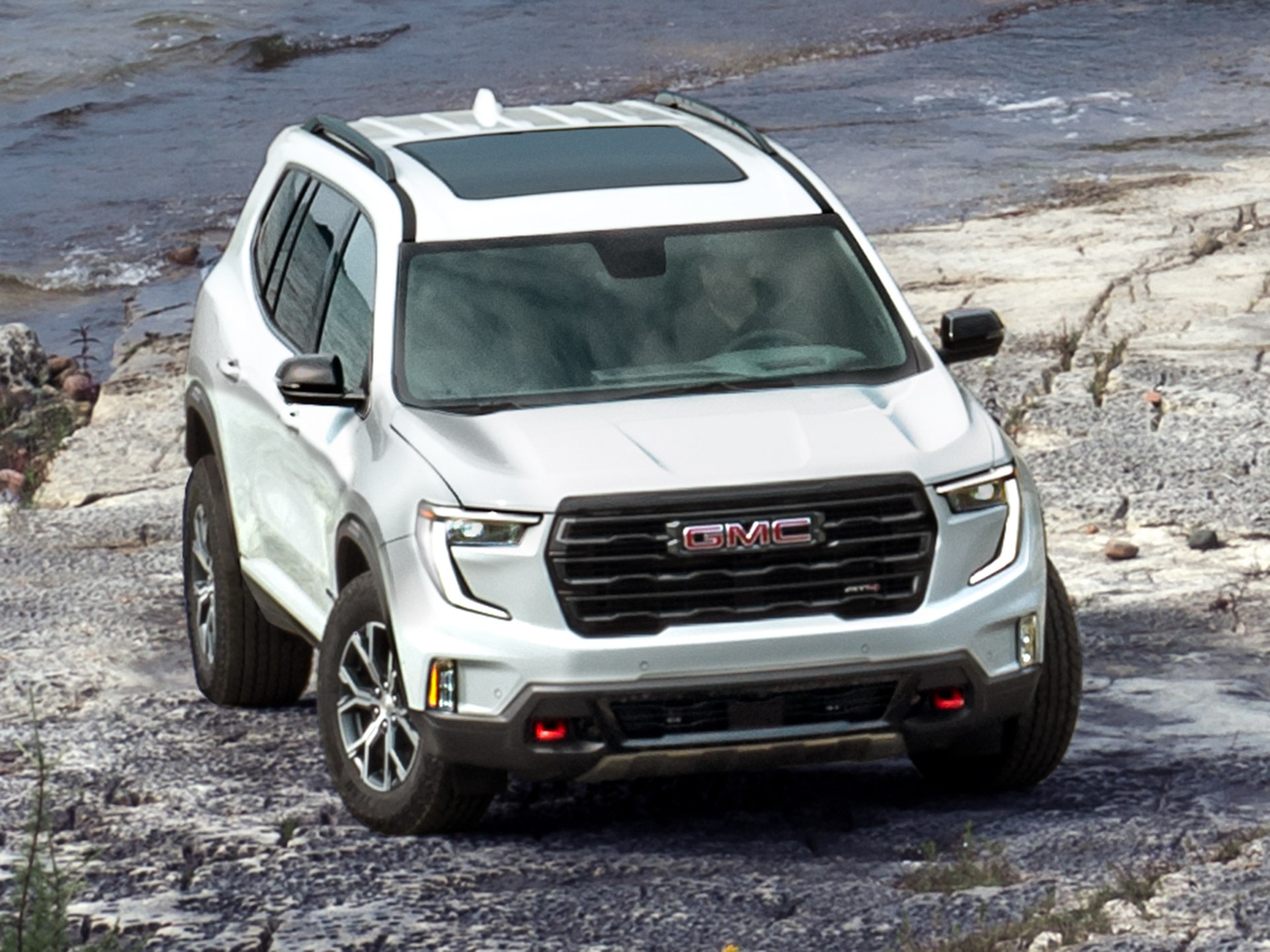Un VUS GMC Acadia blanc 2027 avec toit ouvrant stationné sur un terrain rocheux près de l’eau.