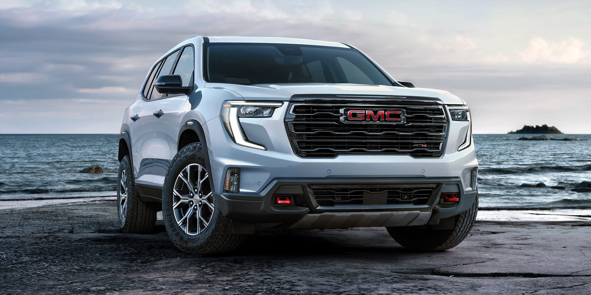 VUS GMC Acadia blanc 2027 avec le logo GMC sur la calandre, stationné sur une plage rocheuse au bord de l’océan.
