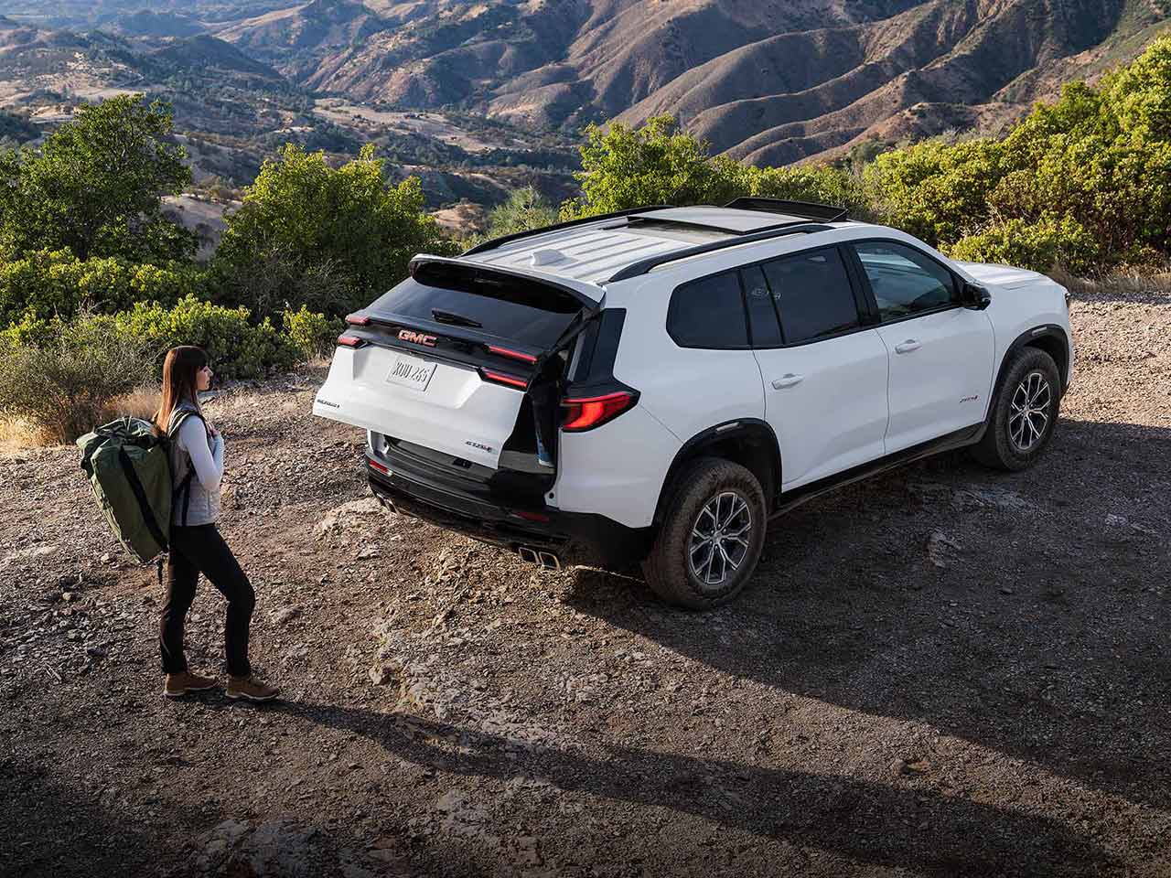 Une femme avec un sac à dos de randonnée, s’approchant d’un VUS GMC Acadia 2027 avec le coffre ouvert.