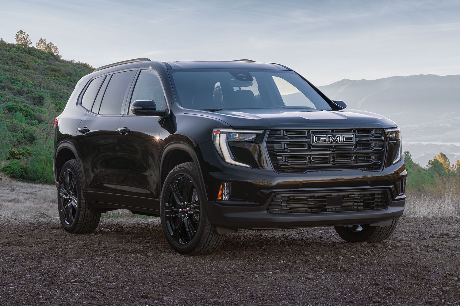 Une GMC Acadia Elevation Black Edition de 2027 garée sur un chemin de terre, avec des collines en arrière-plan et un ciel bleu.