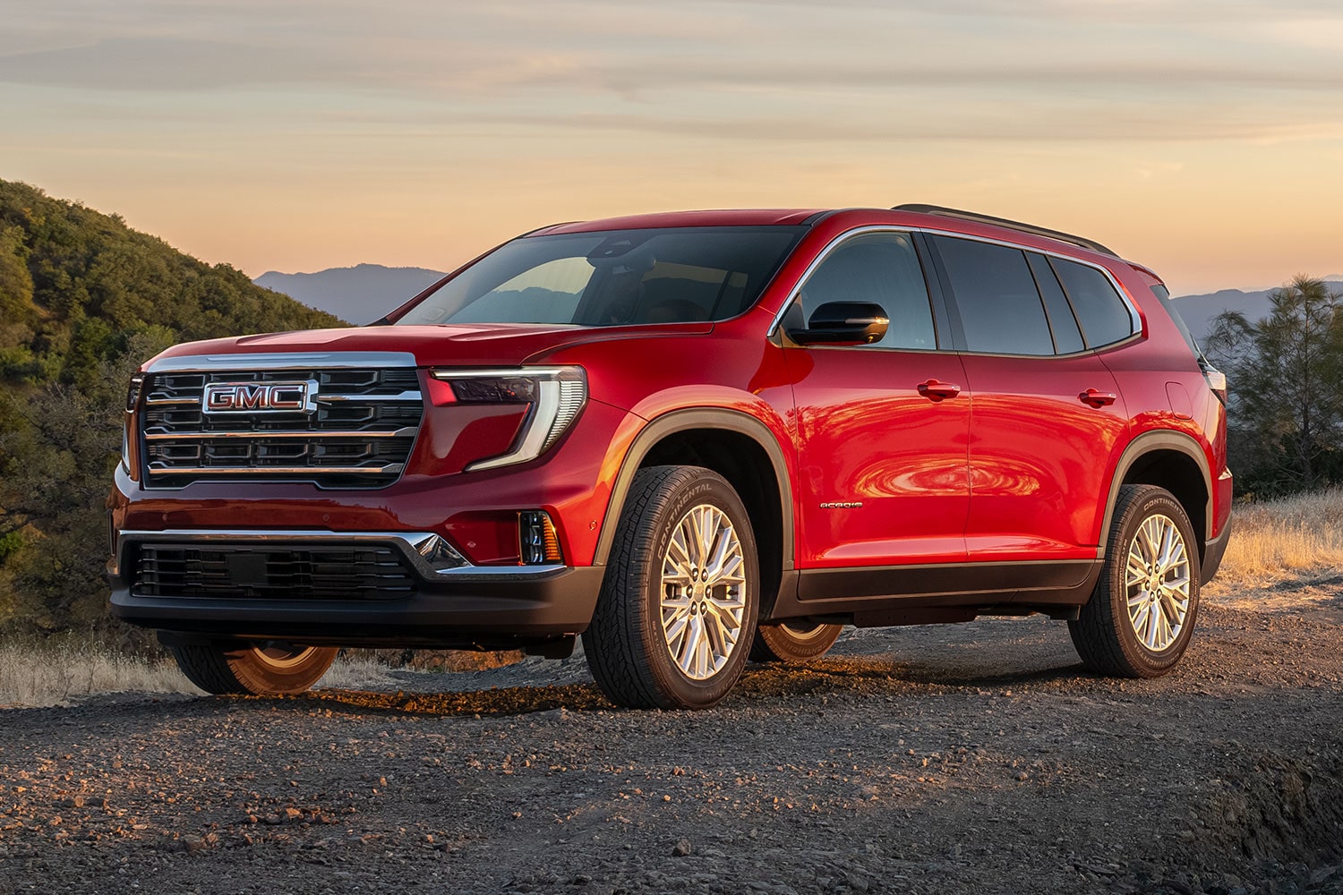 VUS GMC Acadia rouge 2027 stationné sur un chemin de terre au coucher du soleil, dans un paysage vallonné sous un ciel orangé.