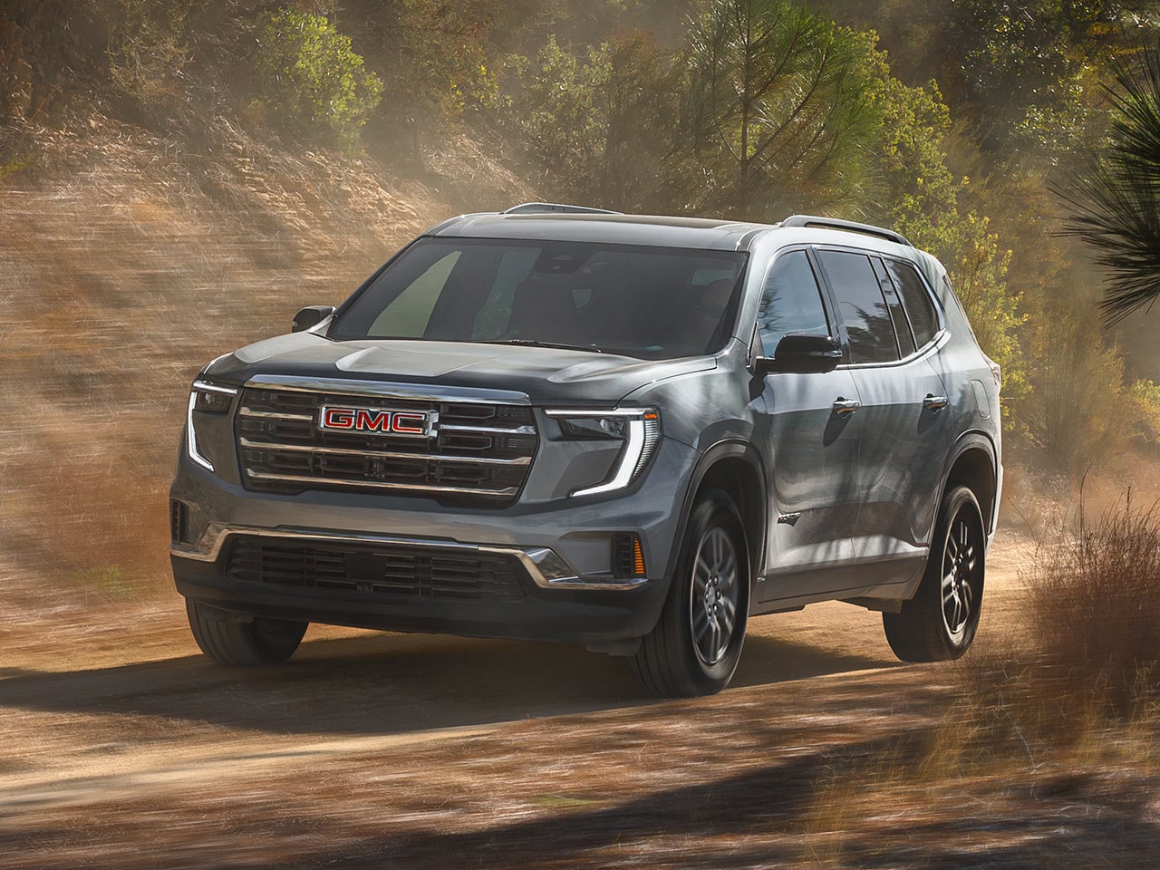 Un VUS GMC Acadia gris 2027 roulant sur une route de terre avec des arbres verts en arrière-plan.