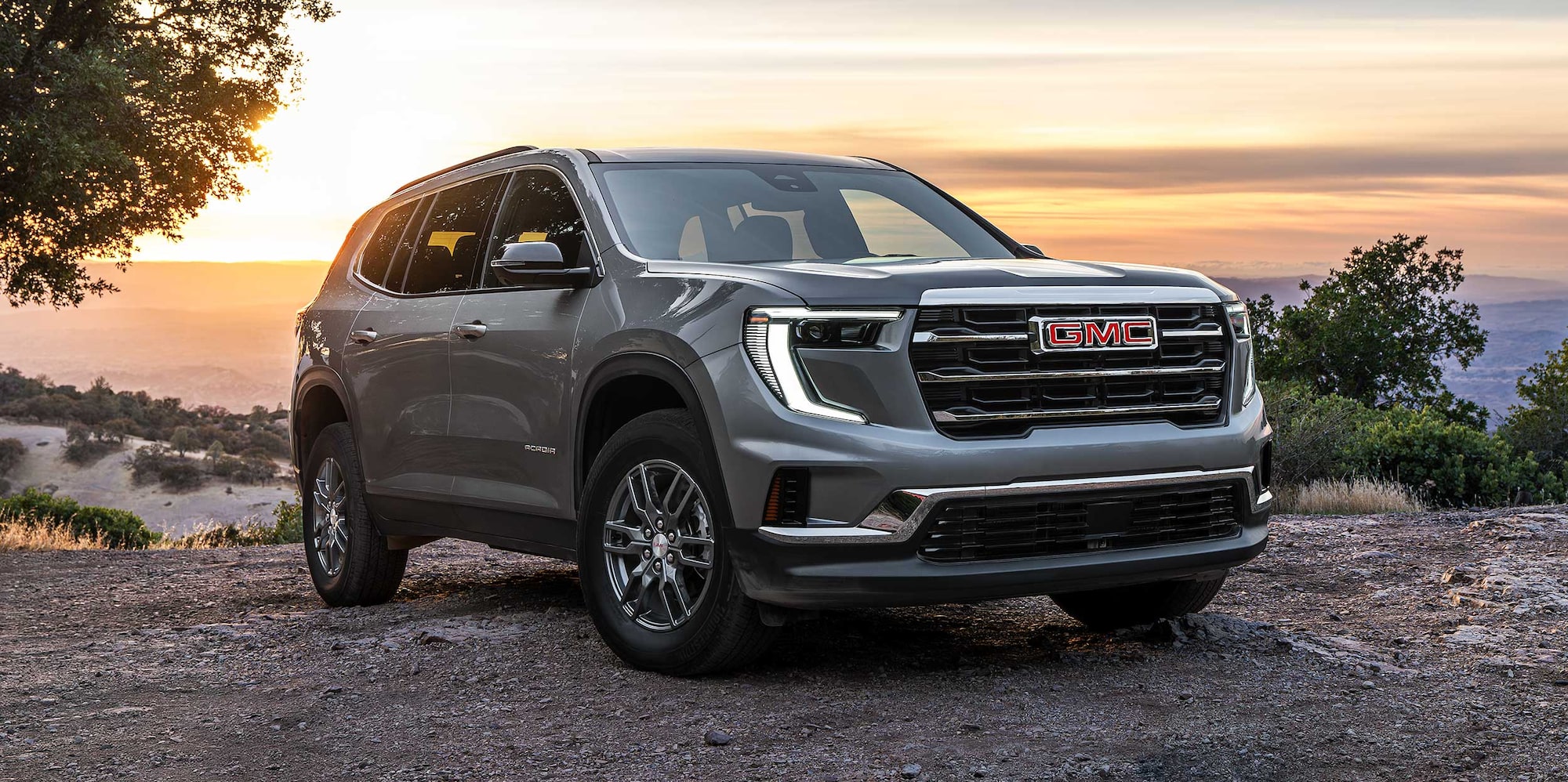 Un VUS GMC Acadia gris 2027 stationné sur une route de terre avec un décor de coucher de soleil et des montagnes au loin.