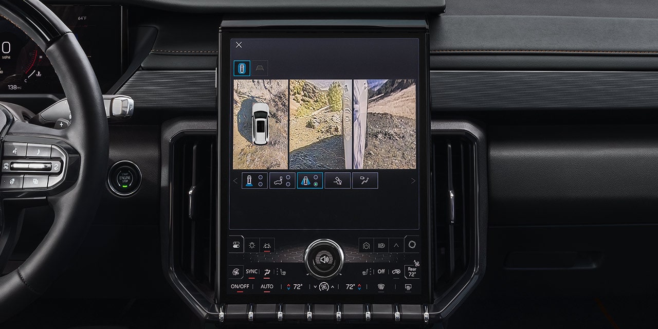 Un gros plan de l’écran d’infodivertissement d’un VUS GMC Acadia 2027 montrant une vue divisée d’un véhicule blanc vu d’en haut et une vue latérale d’une route non pavée.