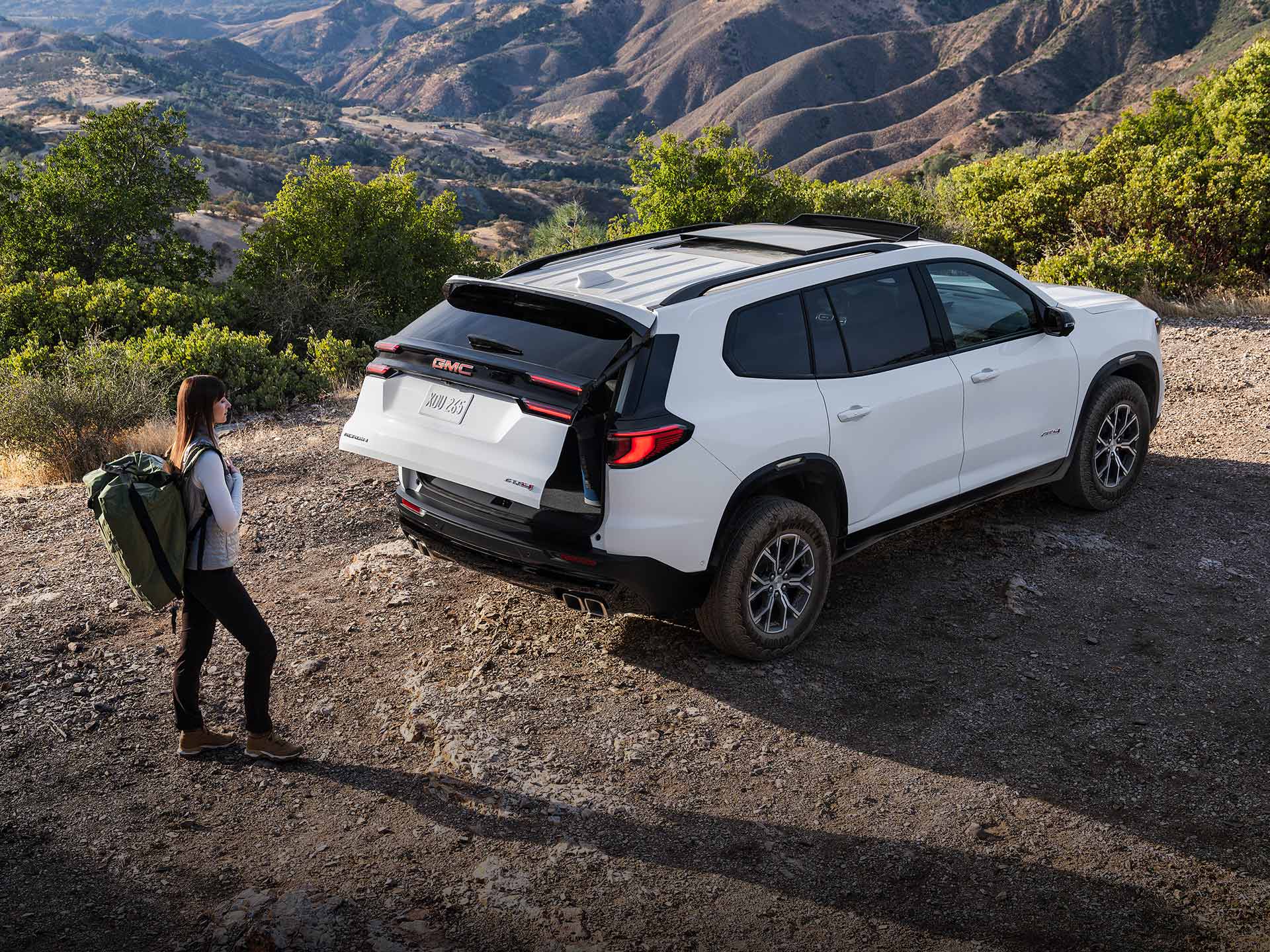 Une femme avec un sac à dos de randonnée, s’approchant d’un VUS GMC Acadia 2027 avec le coffre ouvert.