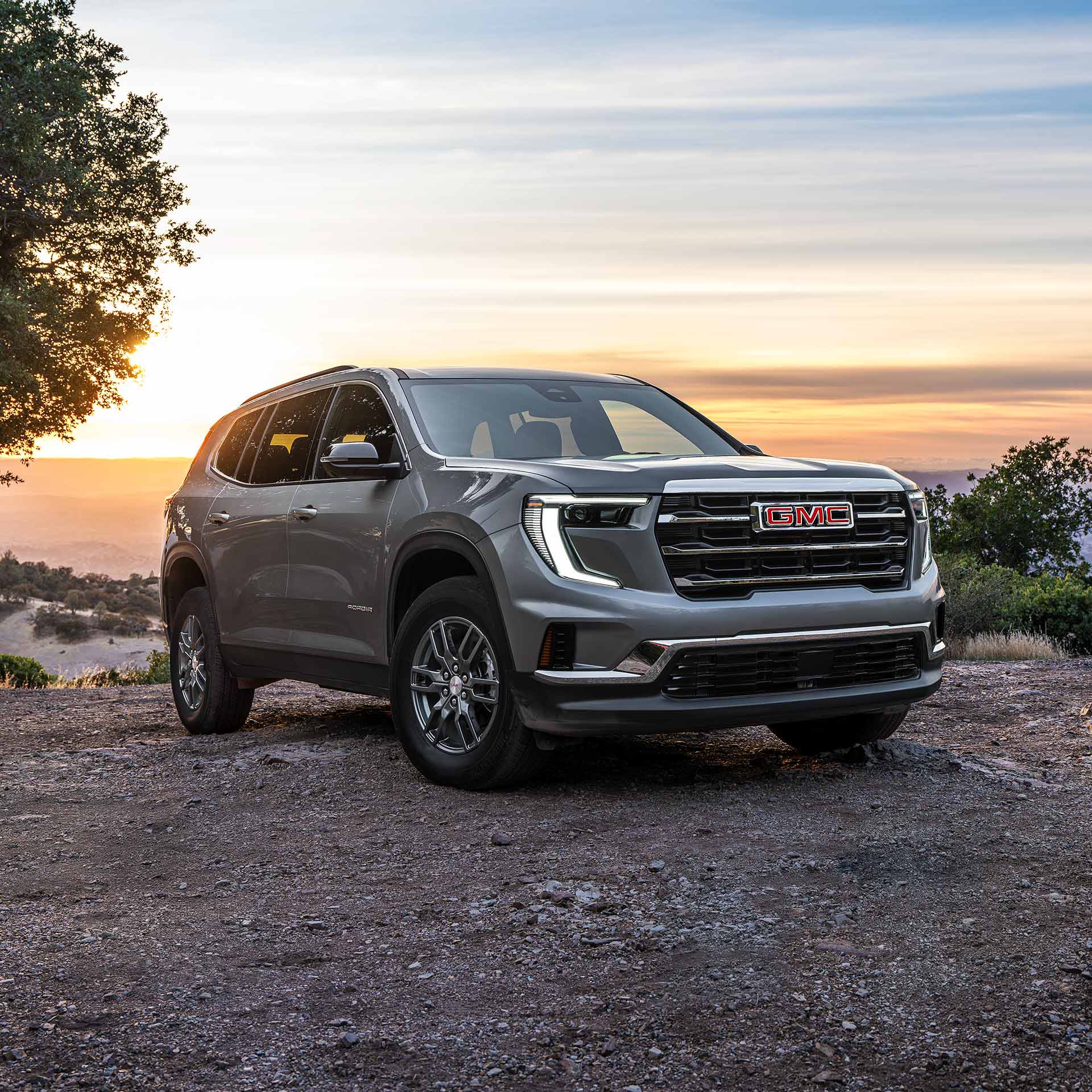 Un VUS GMC Acadia gris 2027 stationné sur une route de terre avec un logo GMC rouge sur la calandre au coucher du soleil.