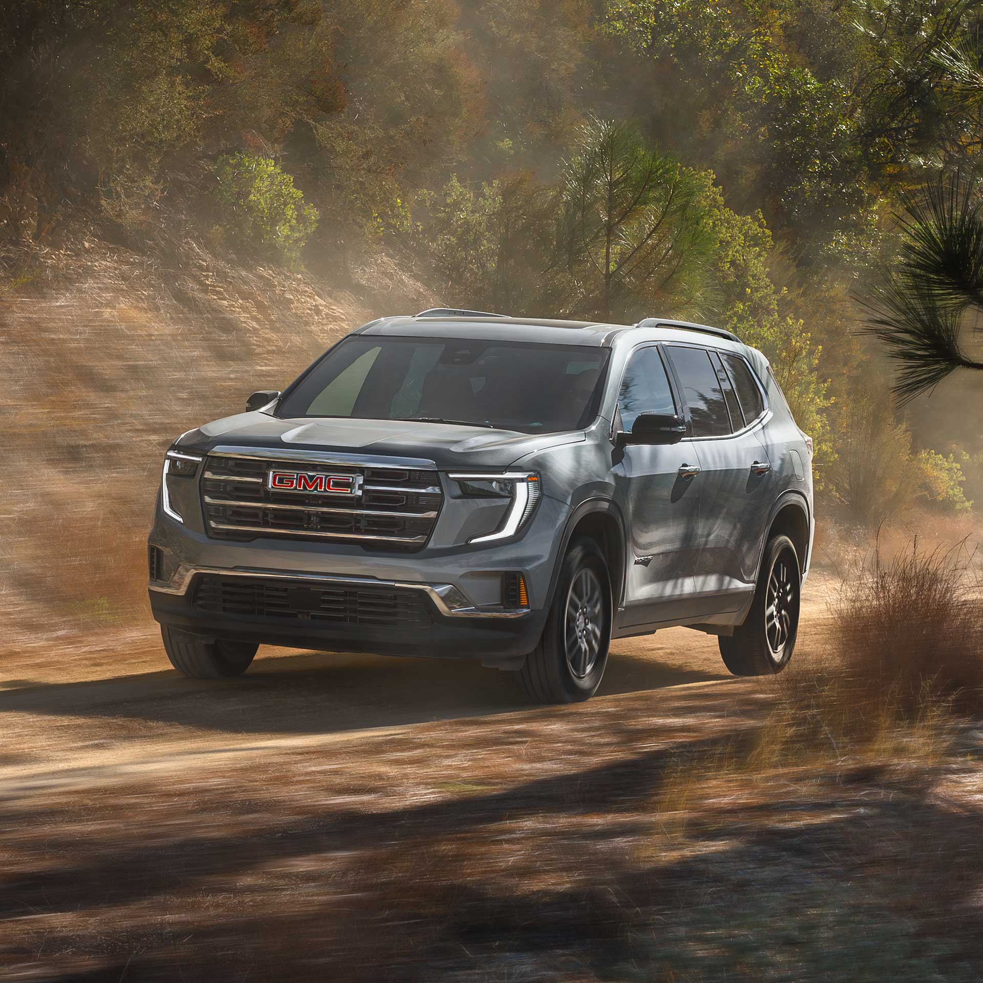 Un VUS GMC Acadia gris 2027 roulant sur une route de terre avec des arbres verts en arrière-plan.