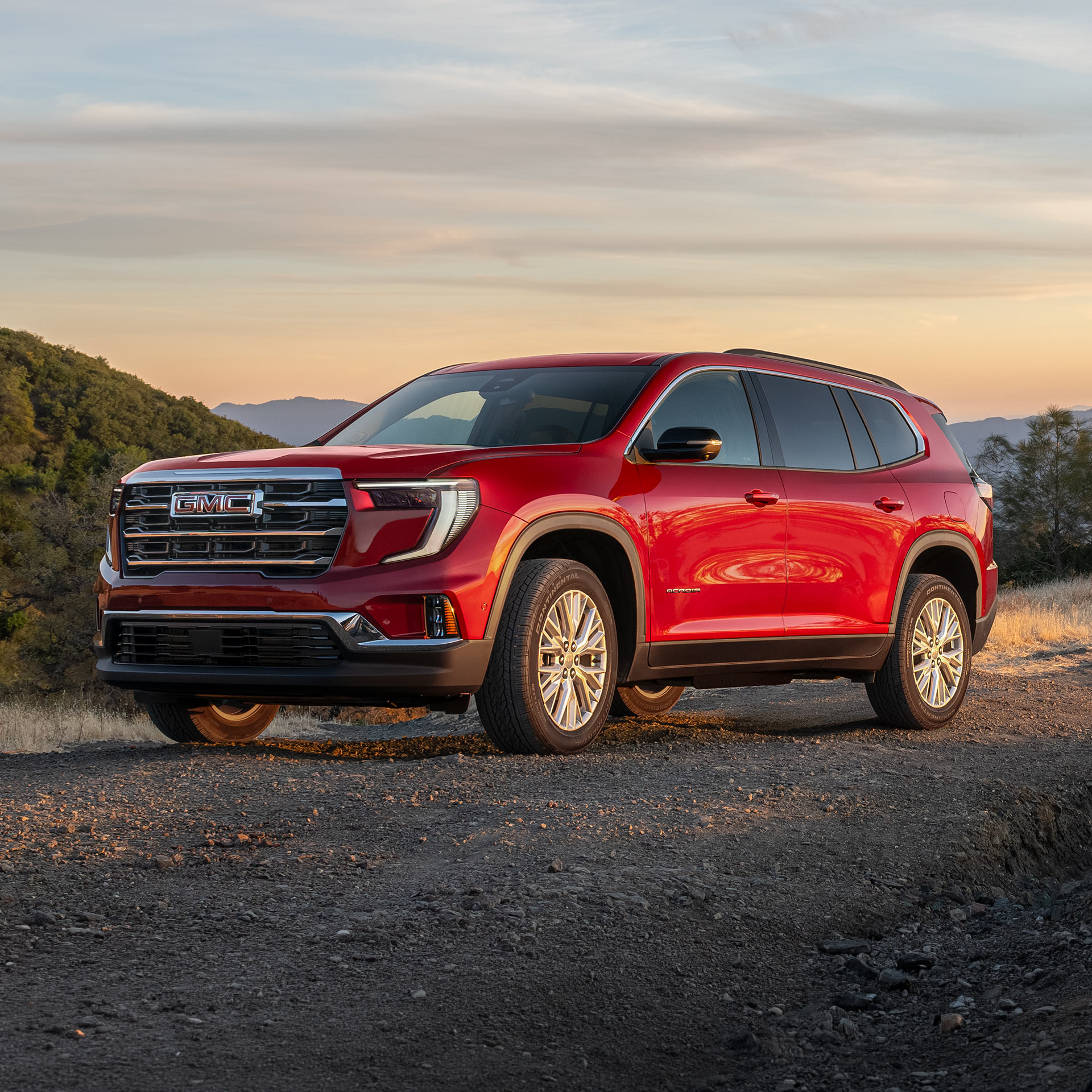 VUS GMC Acadia rouge 2027 stationné sur un chemin de terre au coucher du soleil, dans un paysage vallonné sous un ciel orangé.