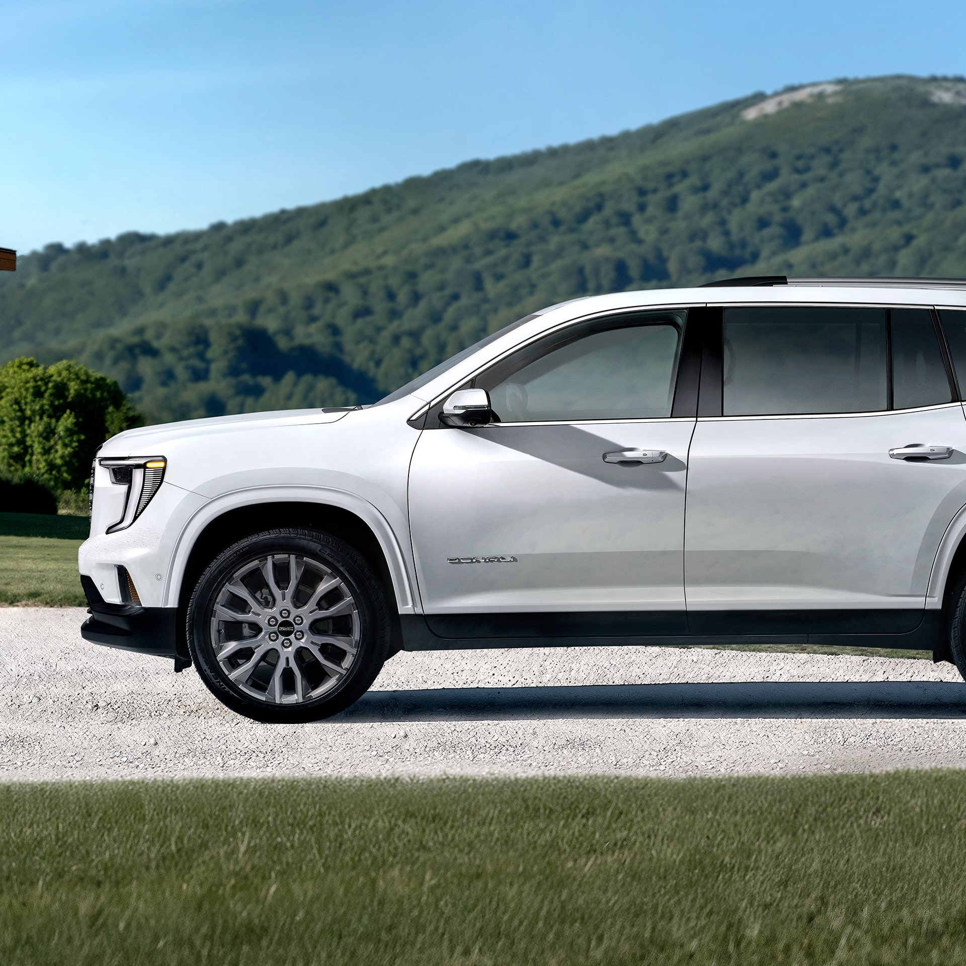 Un VUS blanc GMC Acadia Denali 2027 stationné sur une route de gravier avec des montagnes vertes en arrière-plan.
