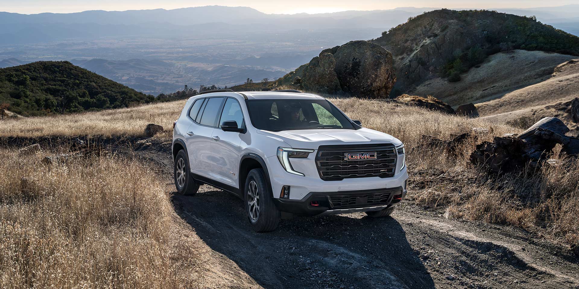 Un VUS GMC Acadia blanc 2027 positionné sur un sentier de terre dans un paysage vallonné et sec, avec des montagnes en arrière-plan.
