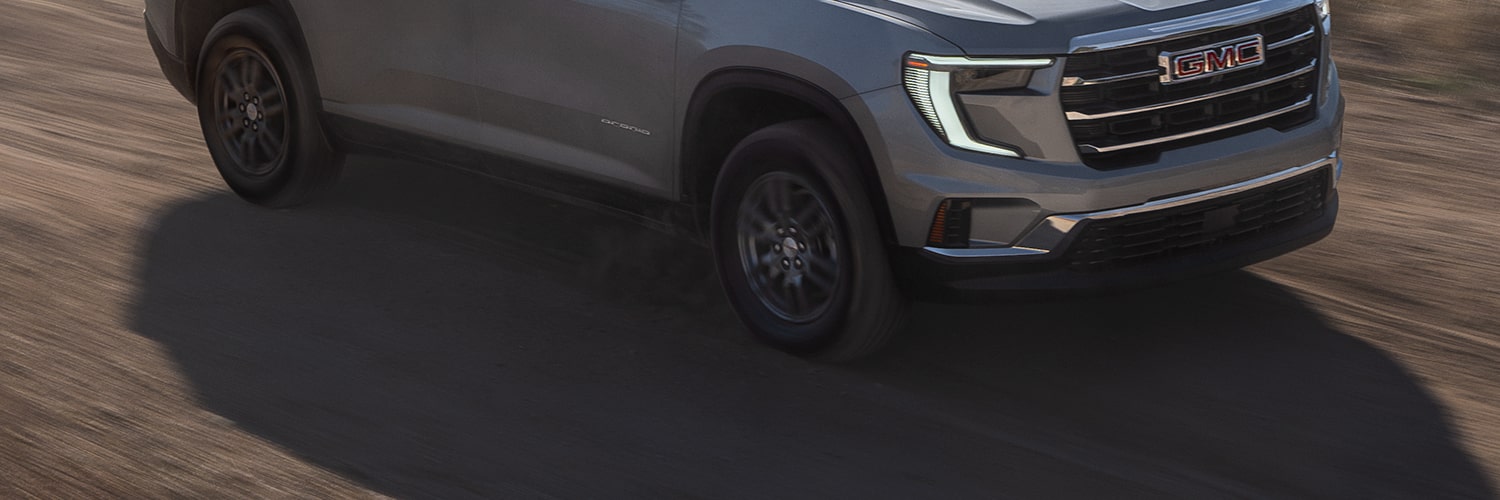 Gray 2027 GMC Acadia SUV Driving on a Winding Dirt Road With Blurred Green Foliage in the Background.