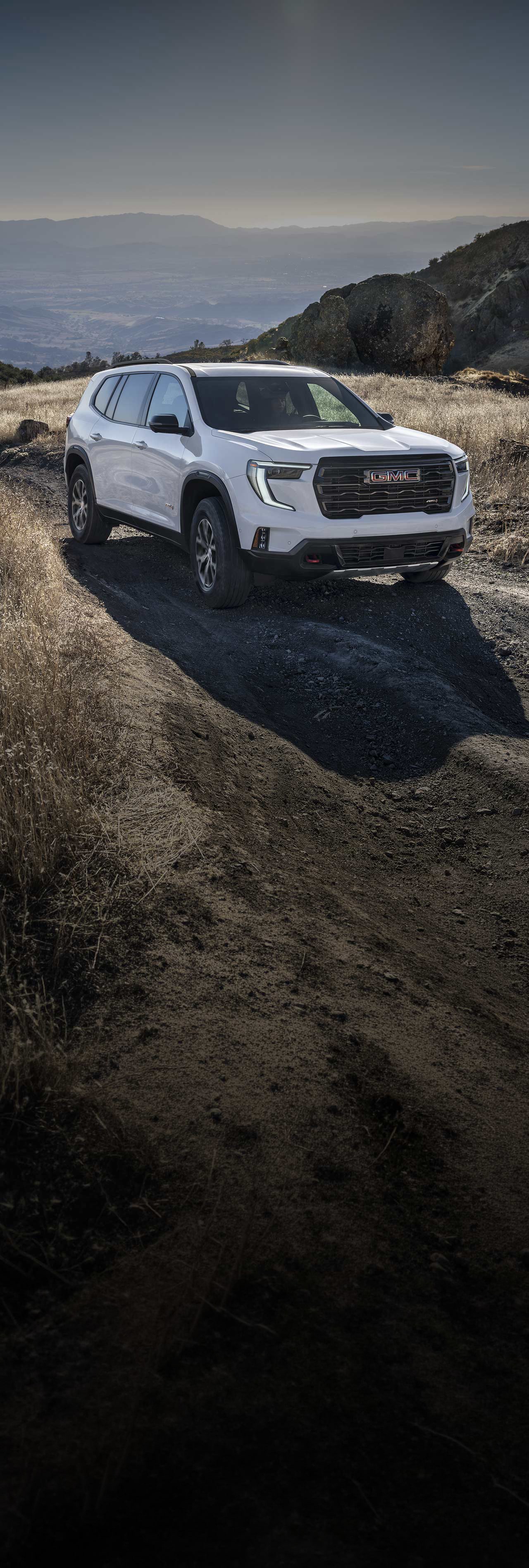 VUS blanc GMC Acadia 2027 sur une route de terre dans un paysage montagneux.