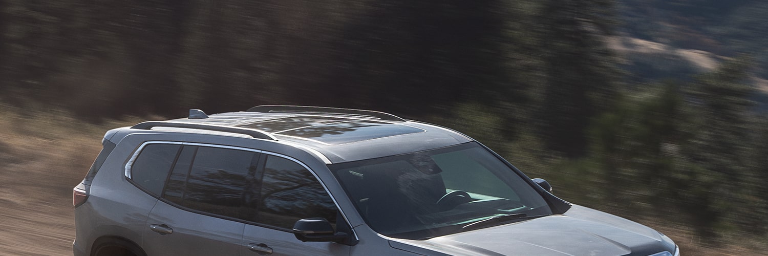 Gray 2027 GMC Acadia SUV Driving on a Winding Dirt Road With Blurred Green Foliage in the Background.