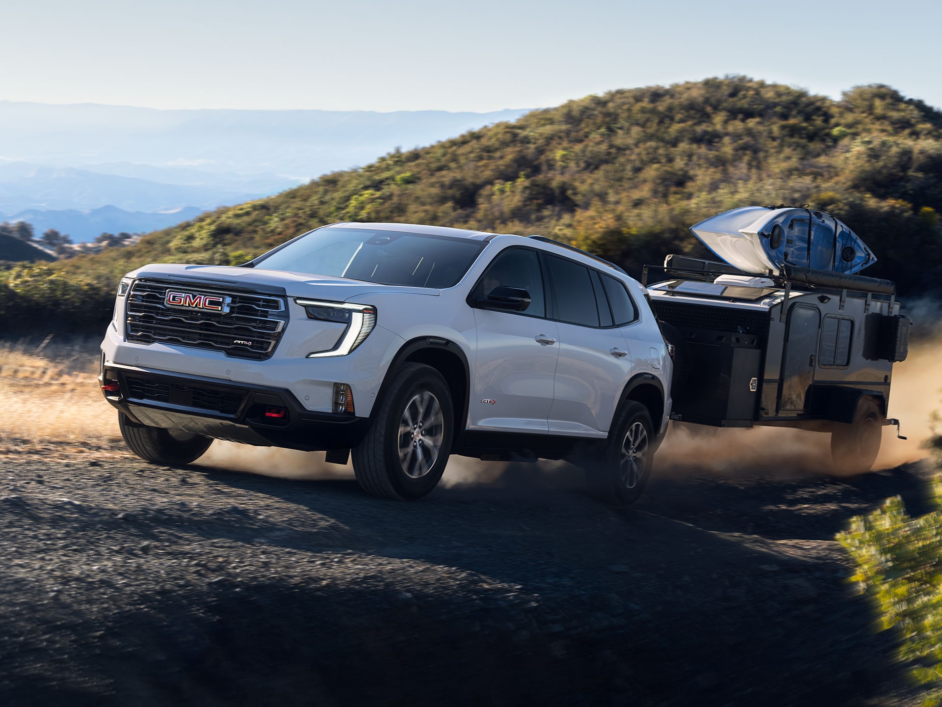 VUS blanc GMC Acadia 2027 tirant une roulotte de camping sombre sur un chemin de terre poussiéreux à travers un paysage vallonné.