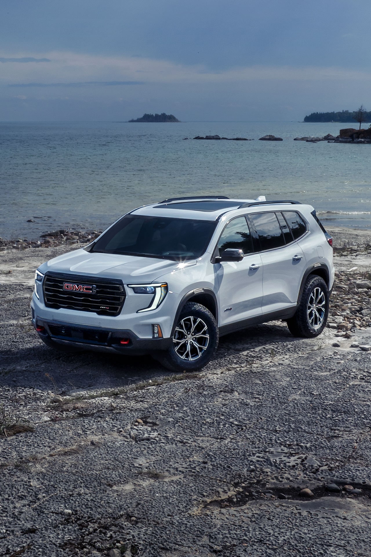 VUS GMC Acadia blanc 2027 avec le logo GMC sur la calandre, stationné sur une plage rocheuse au bord de l’océan.