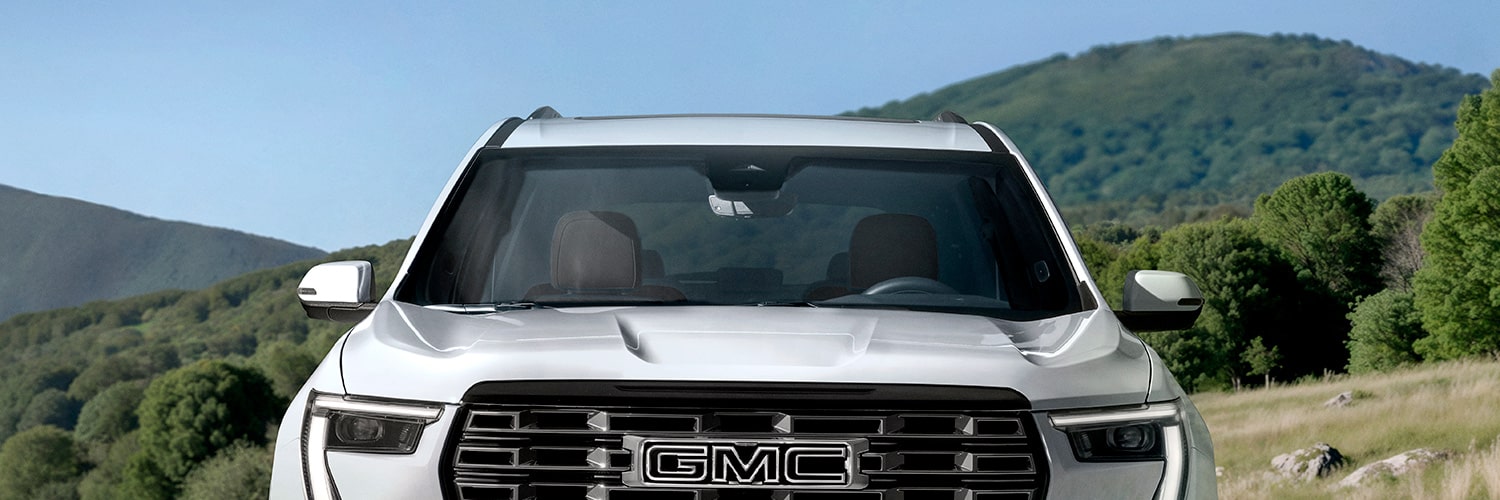 Vue de face d’un VUS GMC Acadia blanc 2027 avec le logo GMC sur la calandre, sur fond de collines ondulantes vertes.