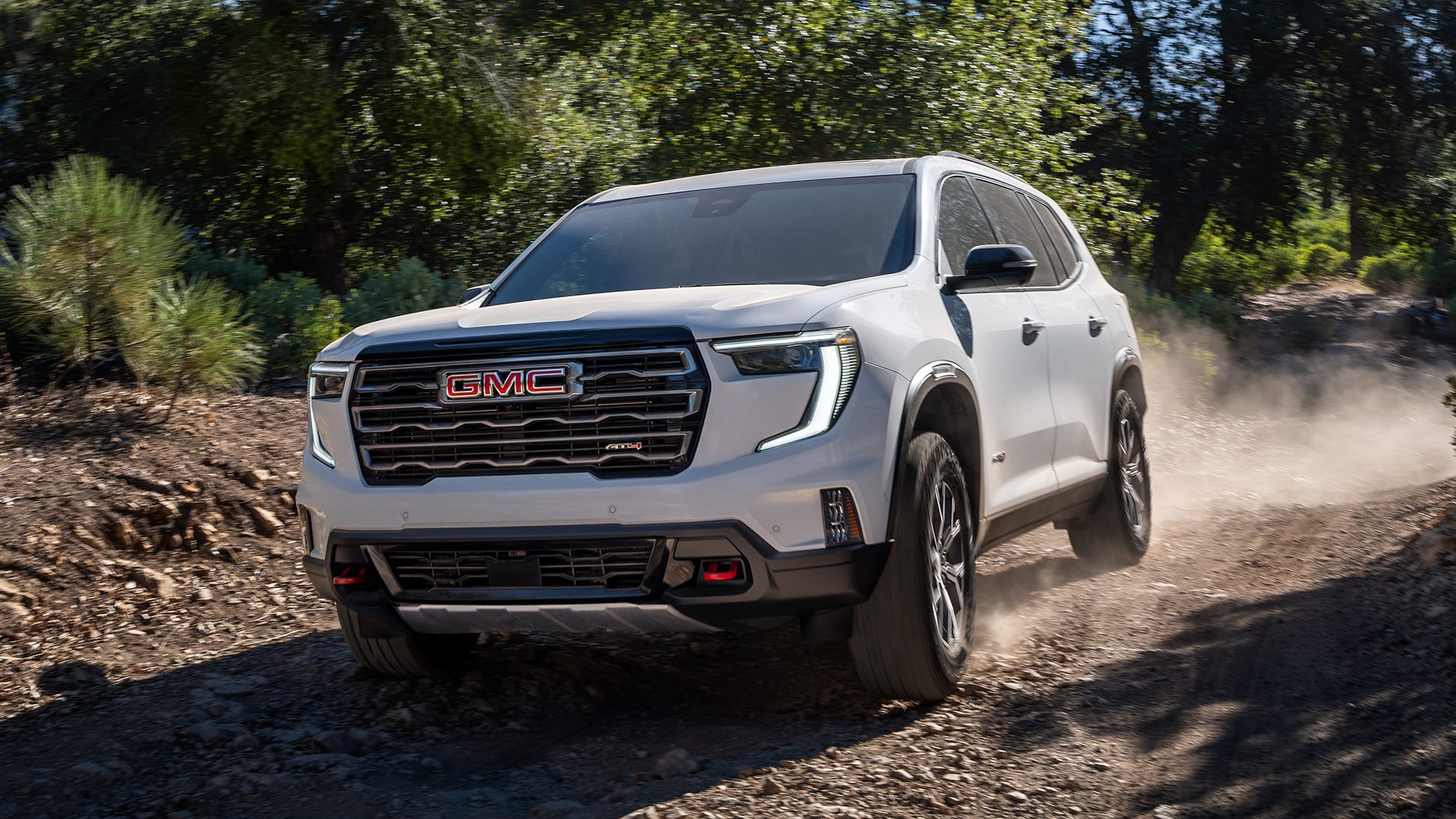 VUS GMC Acadia blanc 2027 roulant sur un chemin de terre poussiéreux à travers une forêt où la lumière du soleil filtre à travers les arbres.