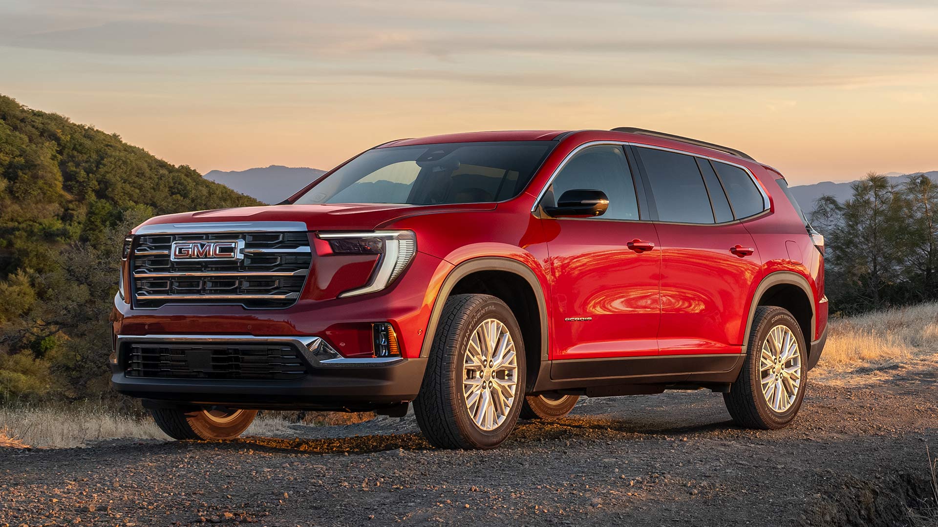 VUS GMC Acadia rouge 2027 stationné sur un chemin de terre au coucher du soleil, dans un paysage vallonné sous un ciel orangé.