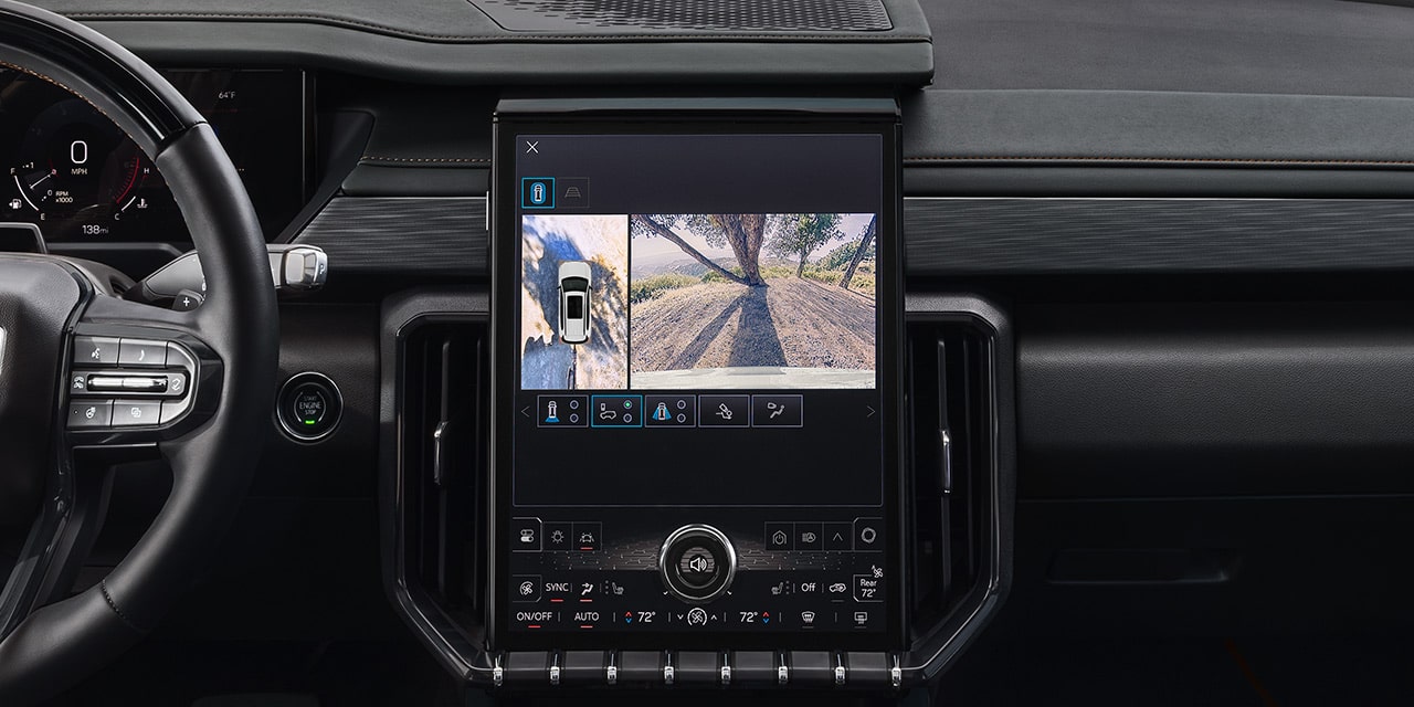 L’écran d’infodivertissement d’un VUS GMC Acadia 2027 affiche une vue divisée d’un VUS blanc vue d’en haut et une vue latérale d’un chemin de terre avec ombres d’arbres.