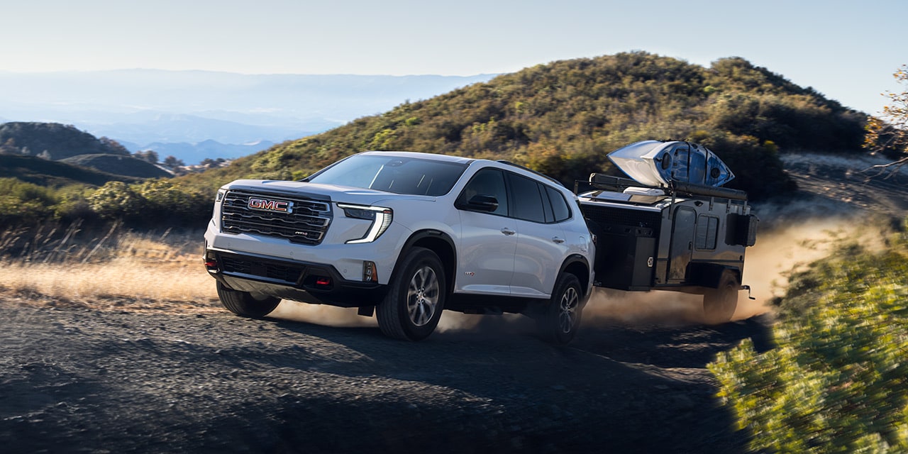 Un VUS GMC Acadia blanc 2027 remorquant une remorque sombre en haut d’une colline poussiéreuse et non pavée, avec des montagnes en arrière-plan sous un ciel dégagé.
