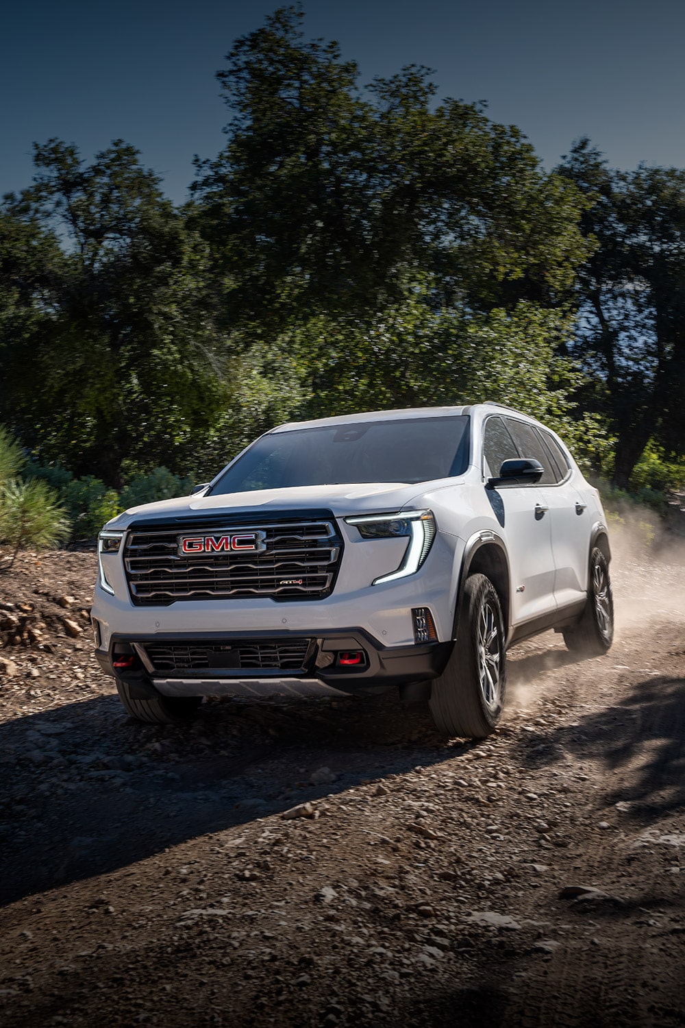 Un VUS GMC Acadia blanc 2027 roulant sur un sentier tout-terrain poussiéreux avec du feuillage vert et des montagnes au loin.