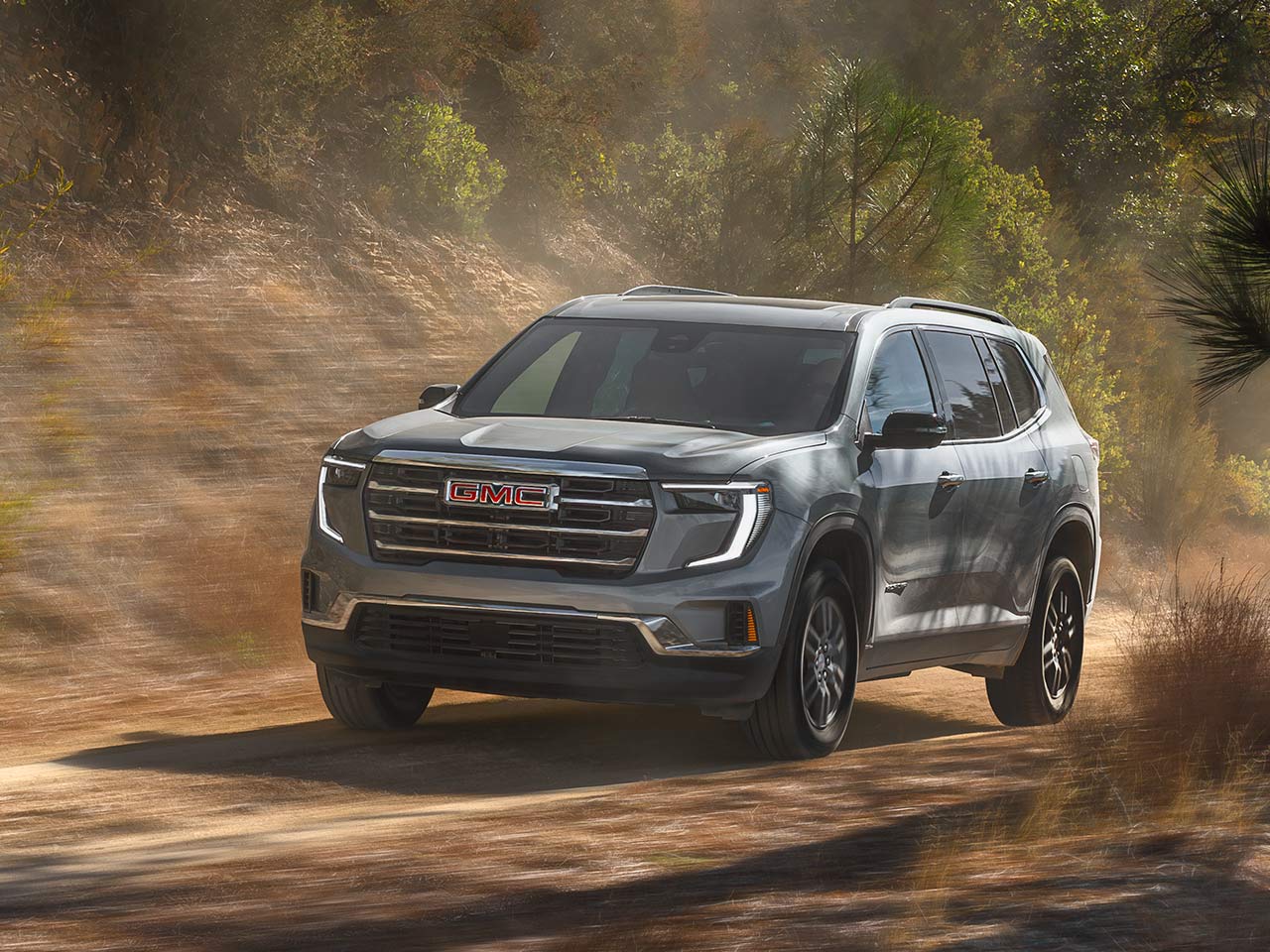 A Gray 2027 GMC Acadia SUV Driving on a Dirt Road with Green Trees in the Background.