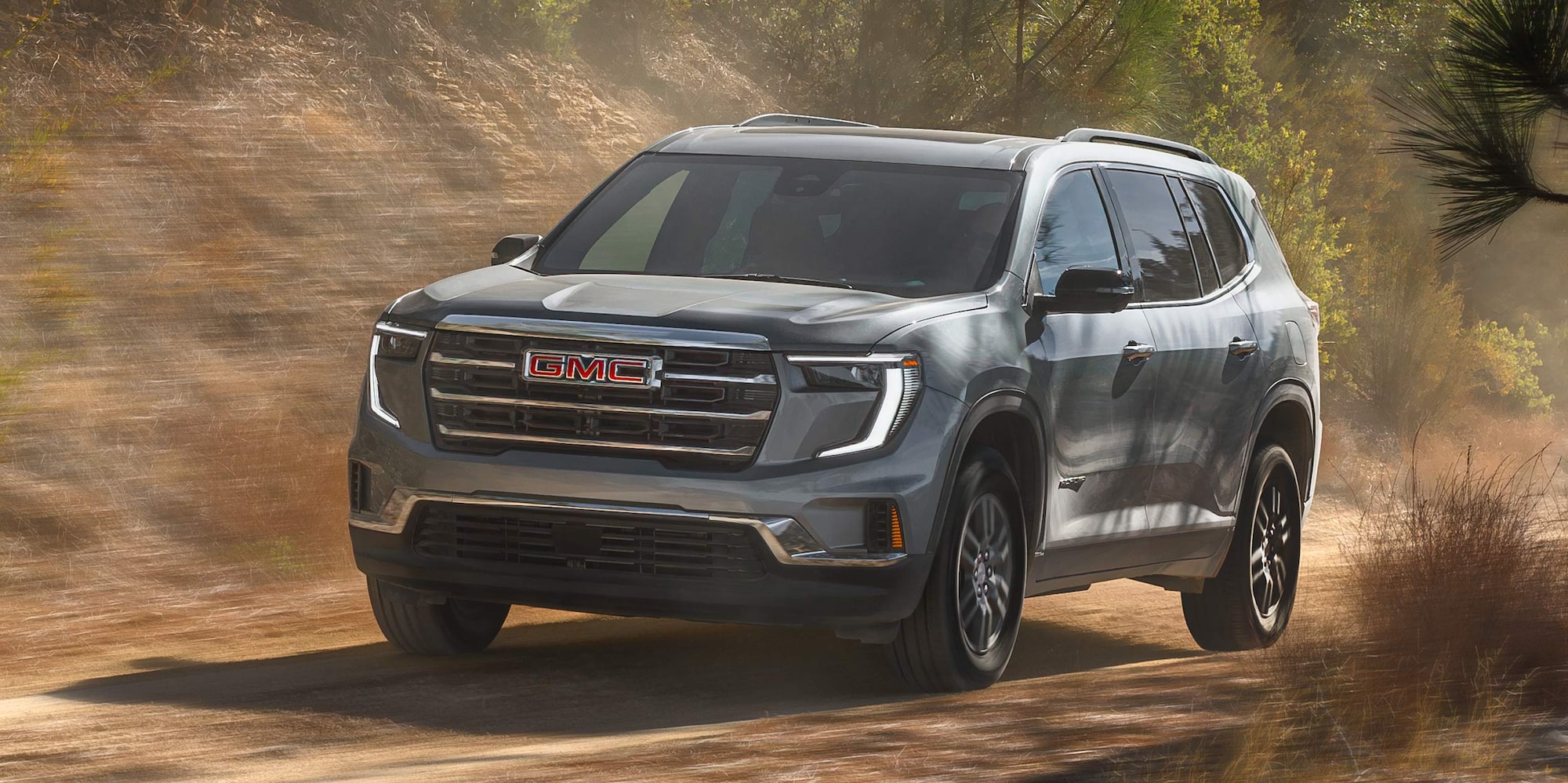 Un VUS GMC Acadia gris 2027 roulant sur une route de terre avec des arbres verts en arrière-plan.