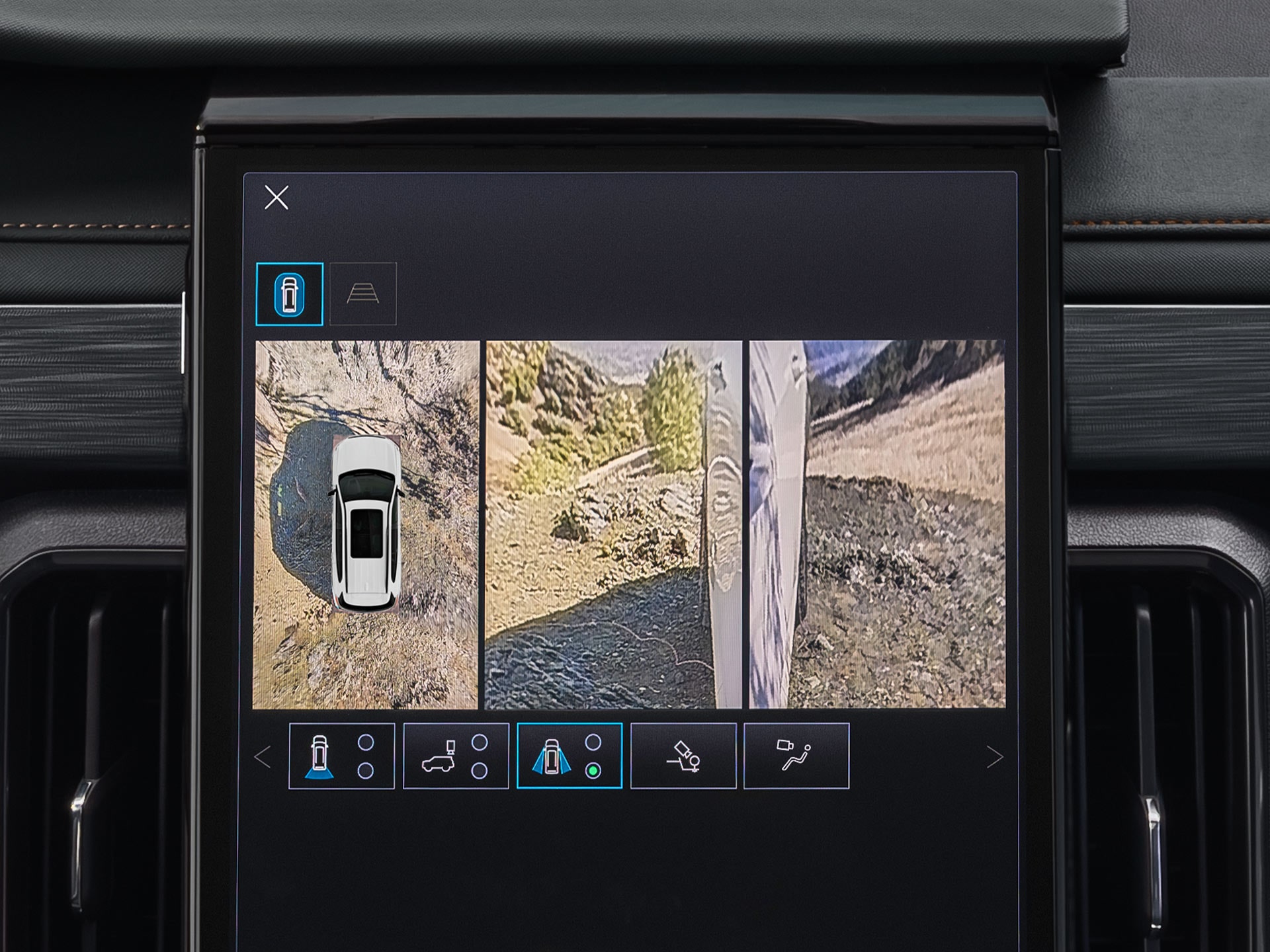 Un gros plan de l’écran d’infodivertissement d’une GMC Acadia 2027 montrant une vue divisée d’un VUS blanc d’en haut, ainsi qu’une vue latérale d’un sentier escarpé et rocheux.