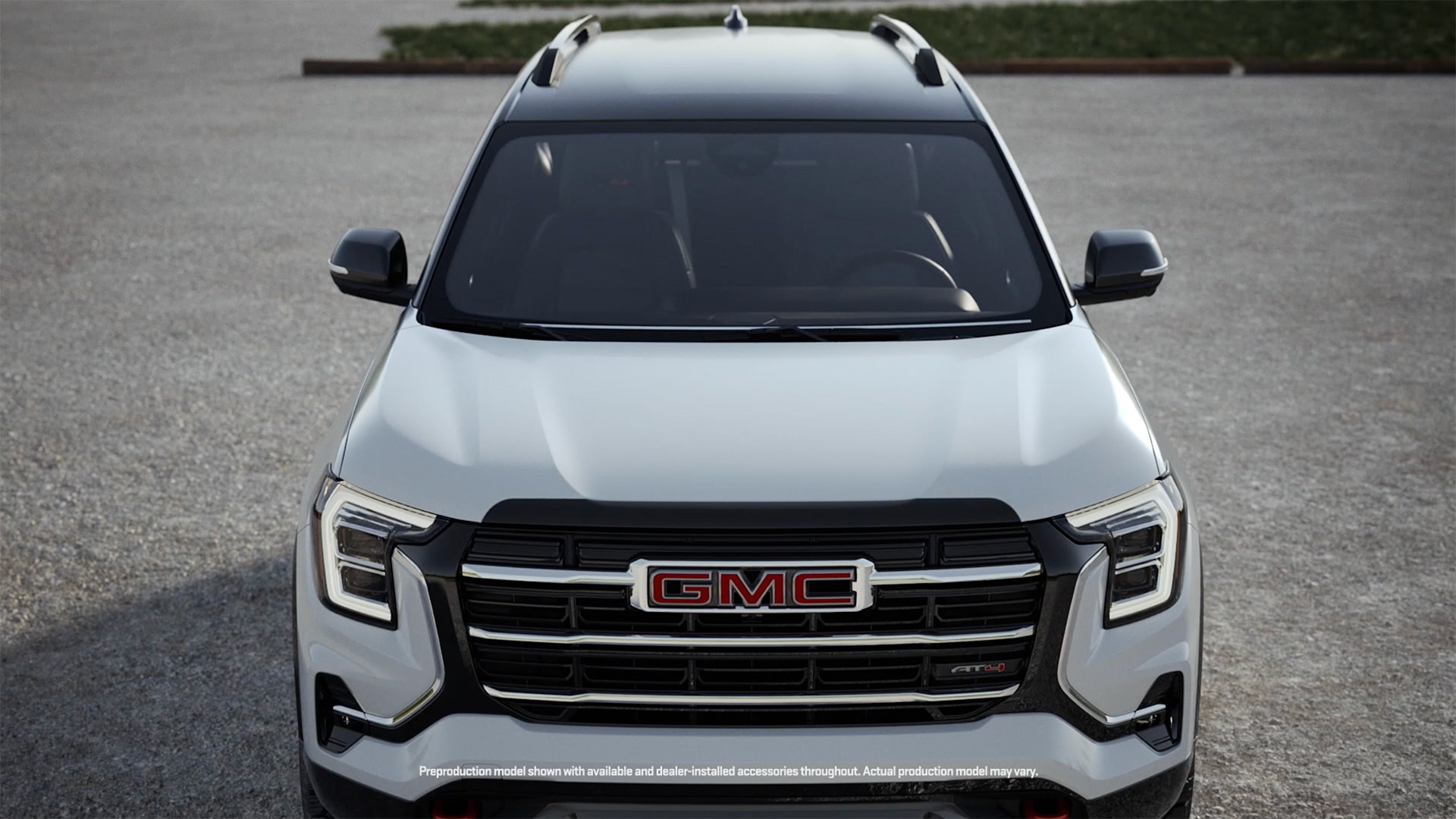 Vue de face d’un SUV AT4 GMC Terrain blanc 2027 avec toit noir, logo GMC Terrain 2027 rouge sur la calandre, phares LED distinctifs et crochets de récupération rouges, garé sur une surface de gravier.