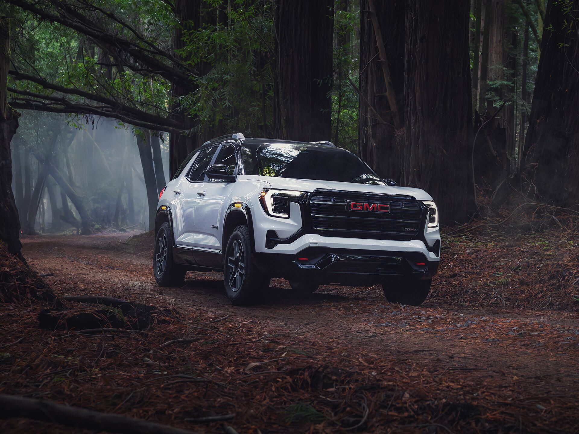 Un SUV GMC Terrain blanc 2027 est garé sur une route de terre au milieu d’une forêt dense aux grands arbres.