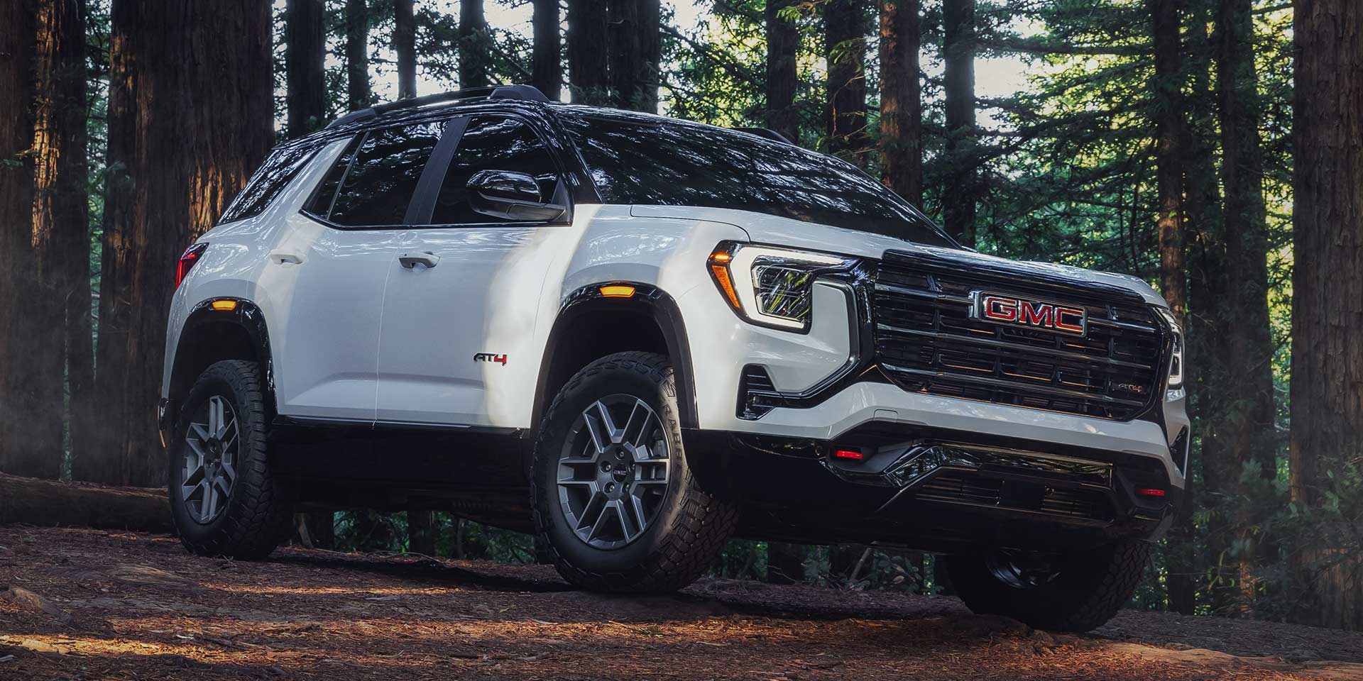 Un SUV AT4 GMC Terrain blanc 2027 avec un toit noir garé sur un chemin de terre forestier entouré de grands arbres.