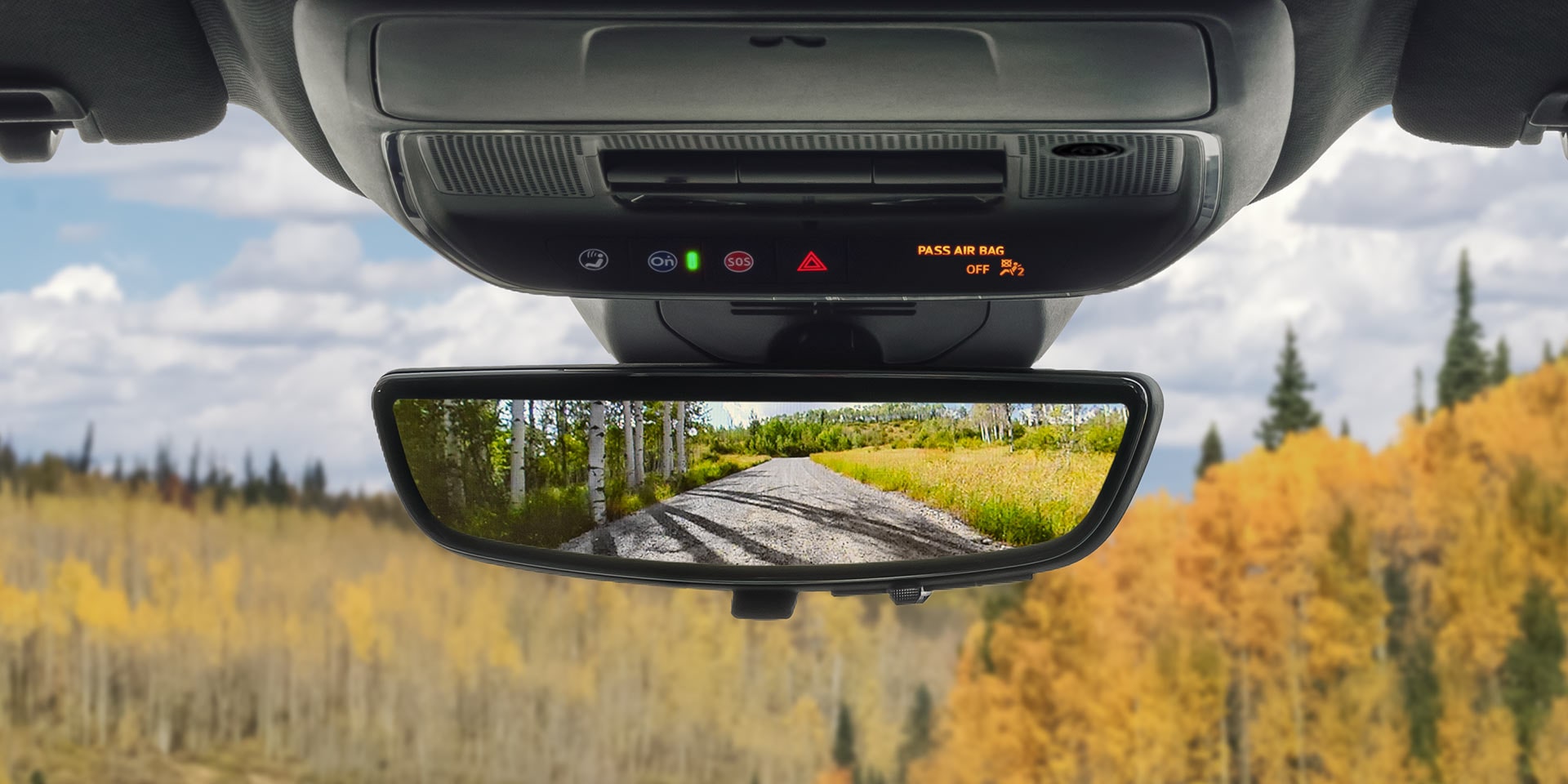 Le rétroviseur d’un véhicule de terrain GMC 2027 reflète une route de terre serpentant à travers des arbres jaunes et verts d’automne.