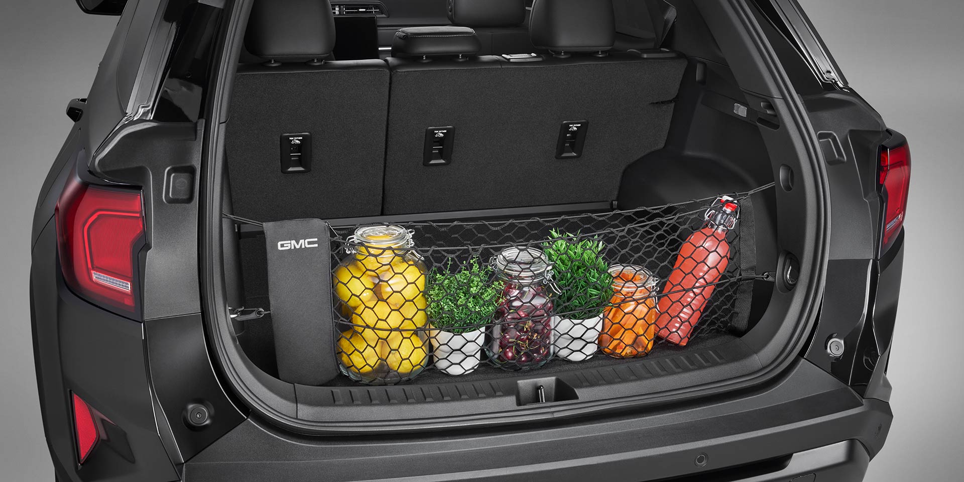 The Open Trunk of a 2027 GMC Terrain Vehicle Showing a Cargo Net Holding Various Items Like Jars of Lemons, Small Plants, and a Red Water Bottle The 2027 GMC Terrain Logo is on the Side of the Trunk.