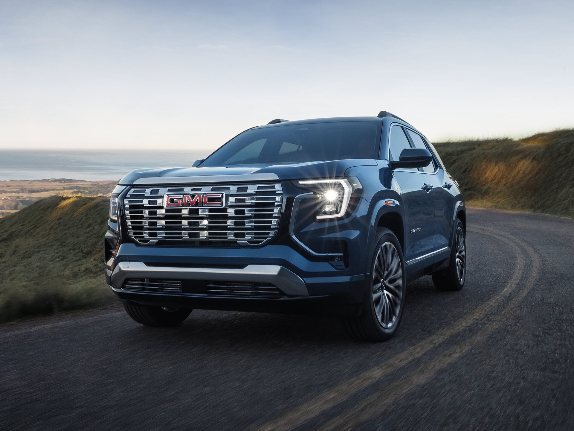A Dark Blue 2027 GMC Terrain Denali SUV Driving on a Winding Road Overlooking a Scenic Coastal Landscape.