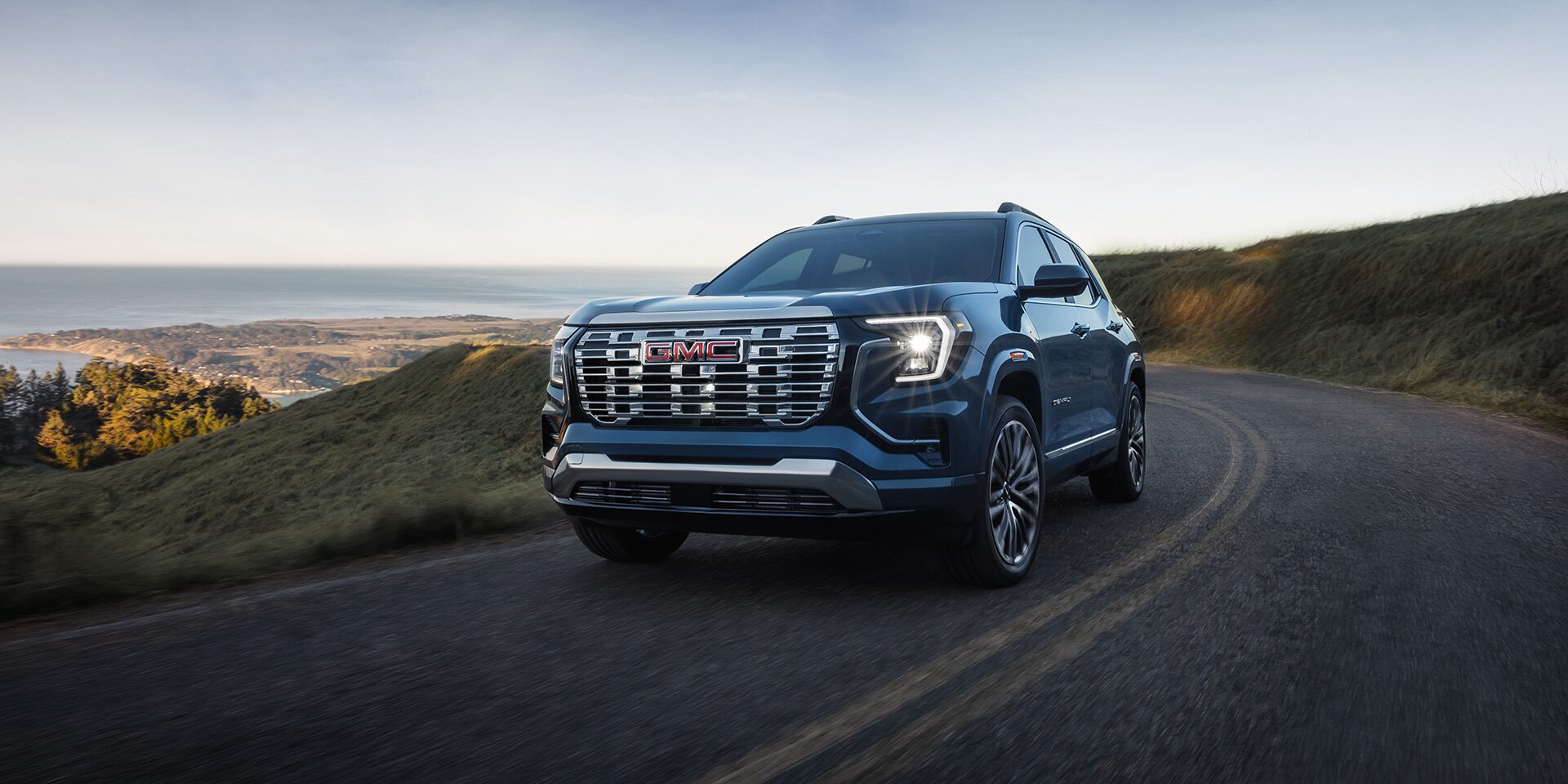 A Dark Blue 2027 GMC Terrain Denali SUV Driving on a Winding Road Overlooking a Scenic Coastal Landscape.