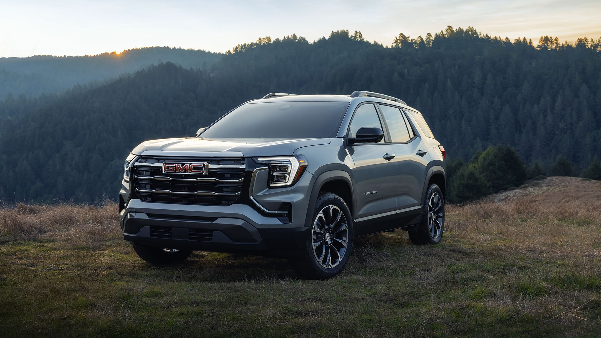 Un SUV gris GMC Terrain Acadia AT4 2027 garé sur une colline herbeuse au coucher du soleil, avec une chaîne de montagnes boisée en arrière-plan.