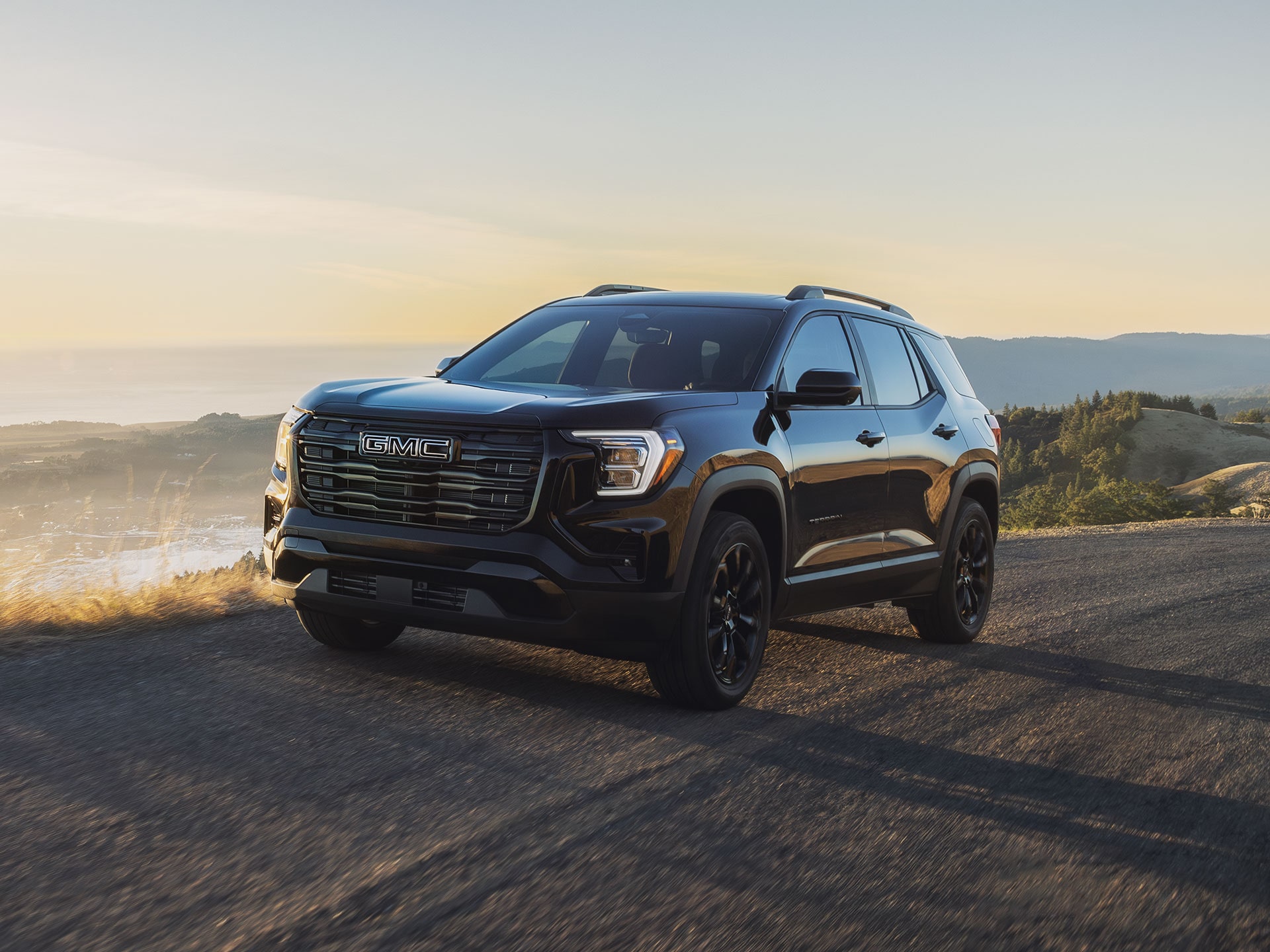 Un SUV terrain GMC noir 2027 roulant sur une route sinueuse avec un soleil d’heure dorée surplombant un vaste océan et un paysage côtier.