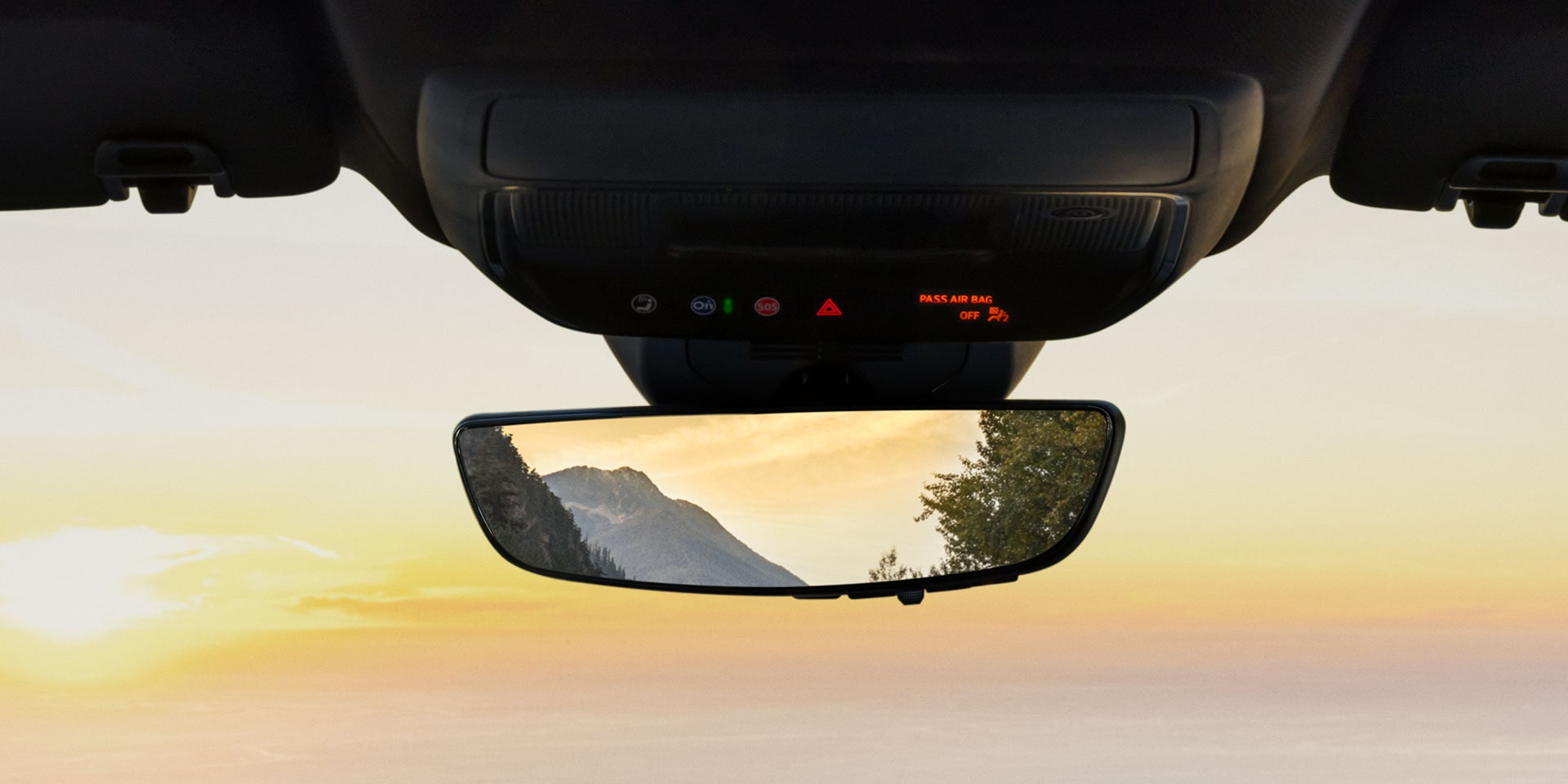Une console intérieure au plafond avec « PASS AIR AIR OFF » affiché au-dessus d’un rétroviseur arrière réfléchissant une montagne et de l’eau au coucher du soleil.
