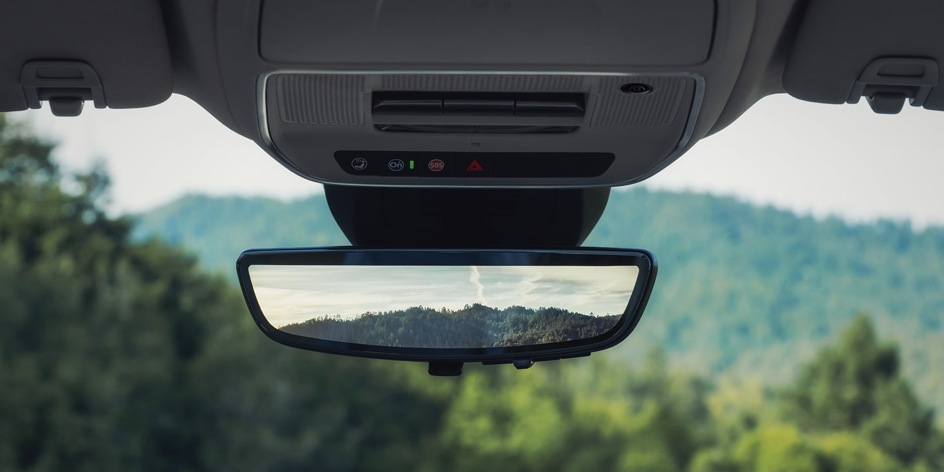 Une console intérieure au plafond avec « PASS AIR AIR OFF » affiché au-dessus d’un rétroviseur arrière réfléchissant une montagne et de l’eau au coucher du soleil.