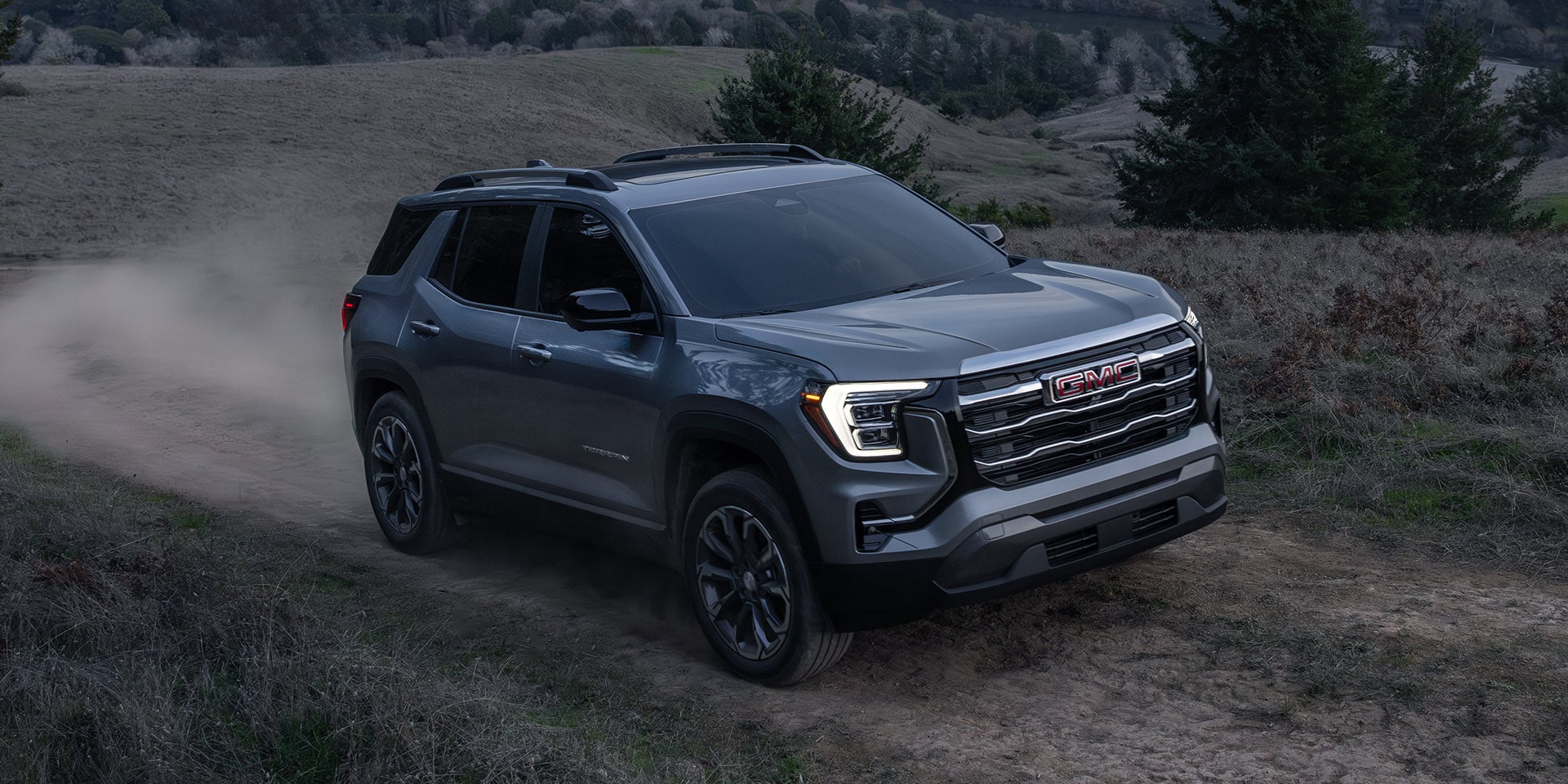Un SUV GMC Terrain Denali gris 2027 soulevant de la poussière alors qu’il roule sur un sentier de terre à travers une vallée.