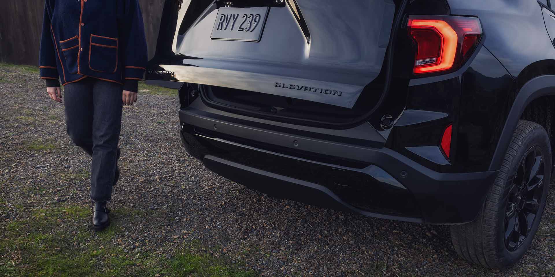 Vue arrière d’un SUV GMC noir 2027 à élévation terrain, avec le hayon ouvert à l’approche d’une personne.